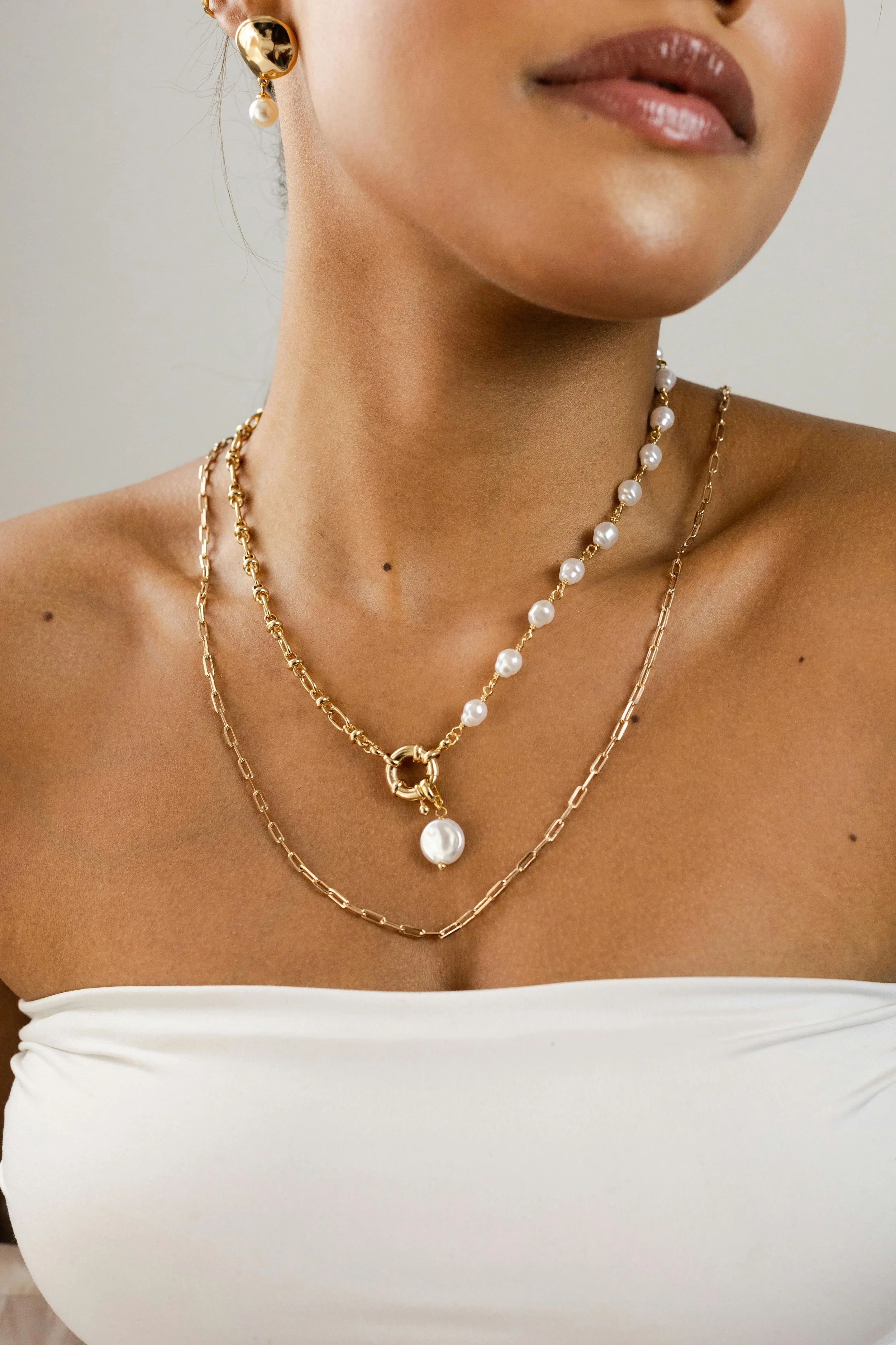 Close-up of a woman wearing gold jewelry, including a pearl earring, two layered gold necklaces with pearls, and a white strapless top.