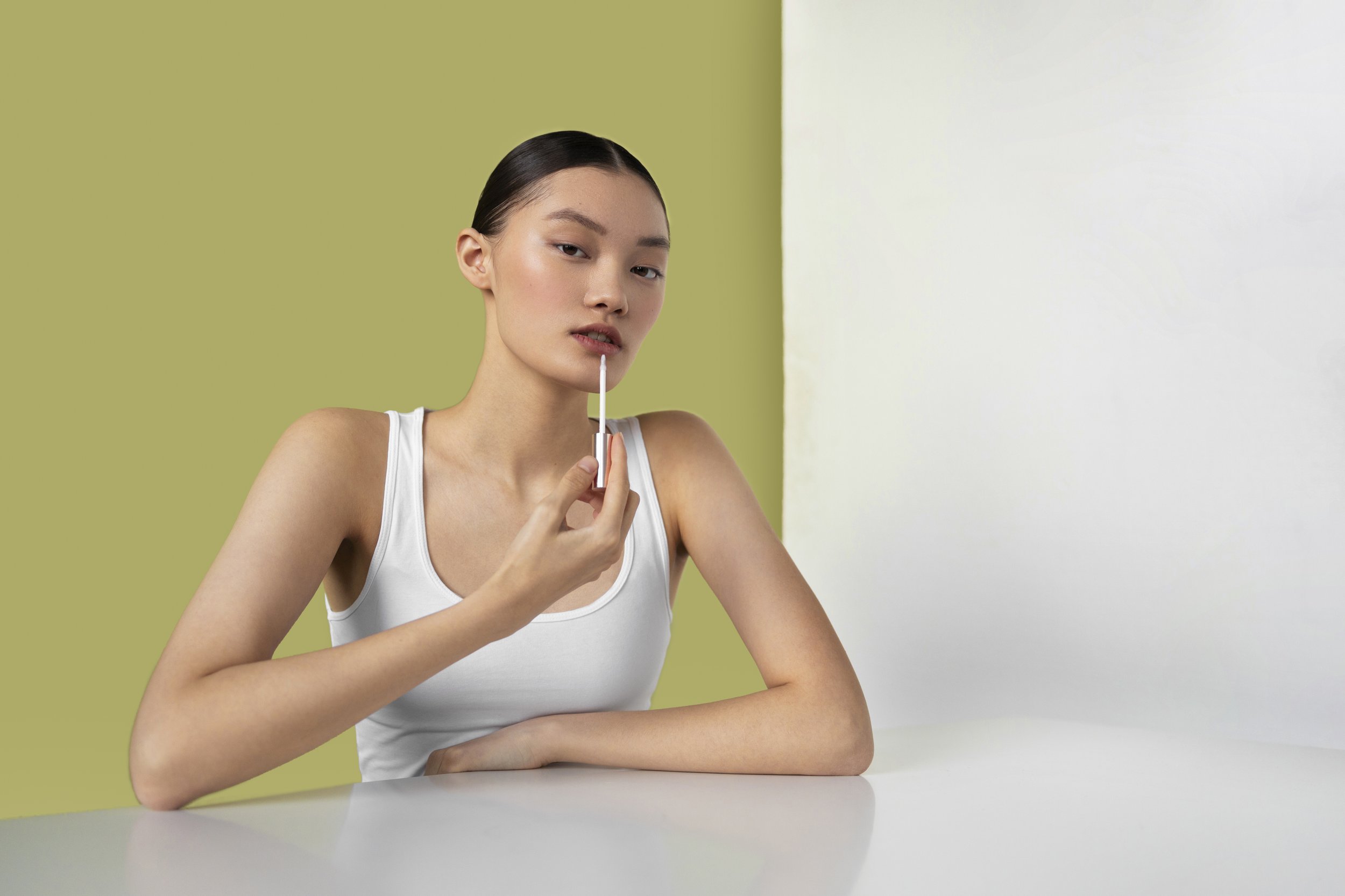 Young woman in white tank top applying lip gloss at white table with pale yellow and white background