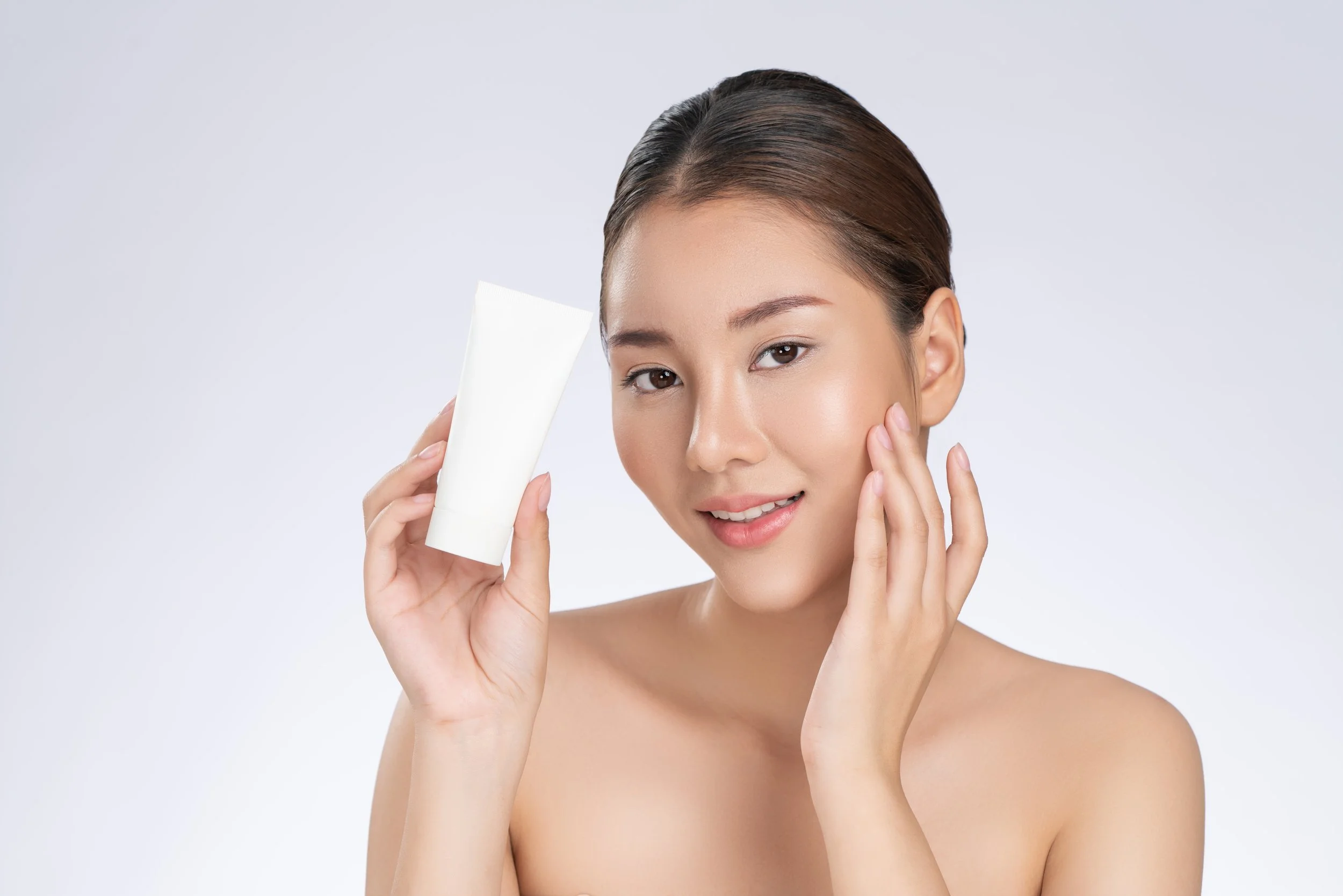 Young woman with clear skin holding a white skincare product tube near her face, smiling softly against a light gray background.