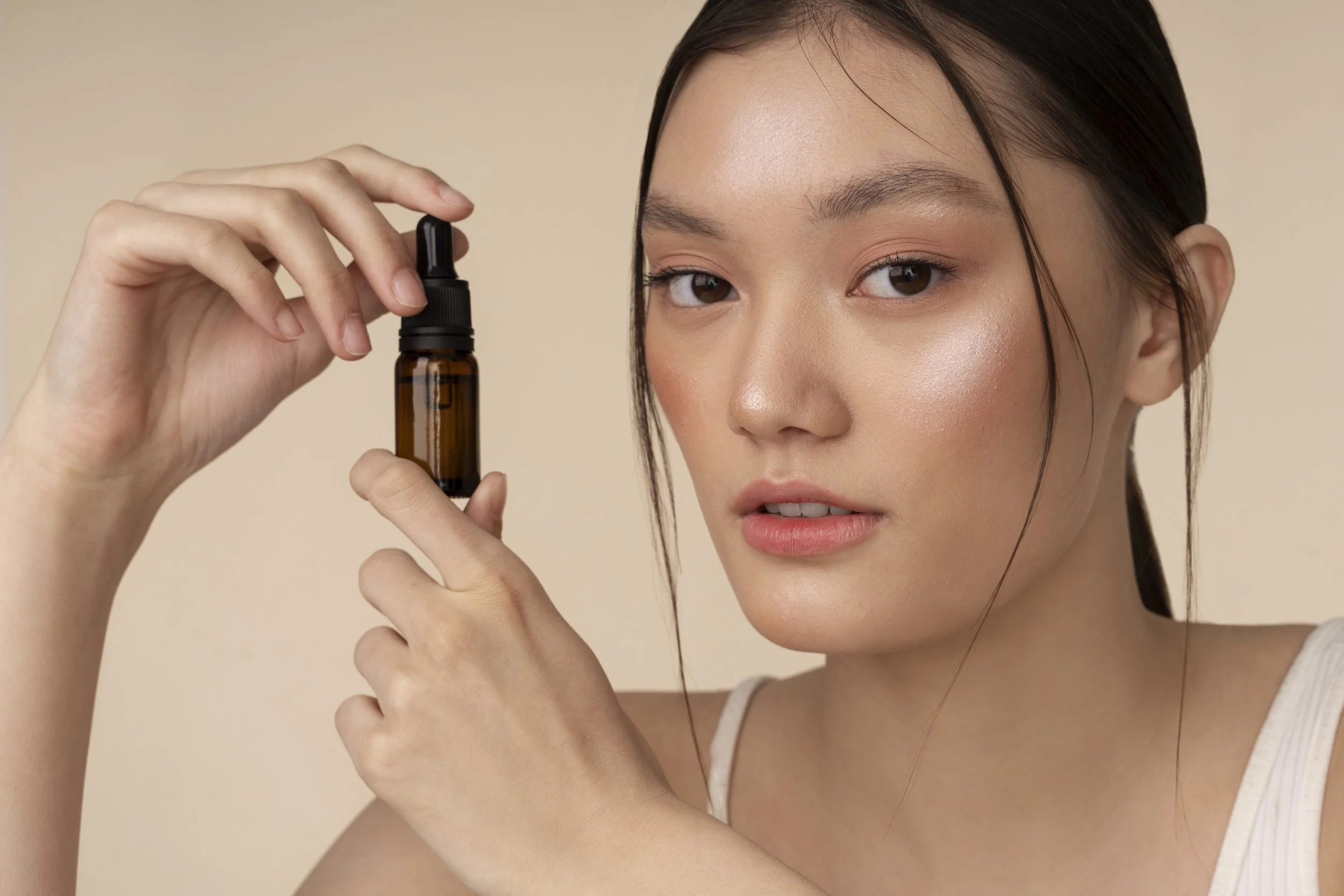 A woman with fair skin and dark hair holds a small amber glass bottle with a black spray top close to her face, looking at the camera with a neutral expression against a beige background.