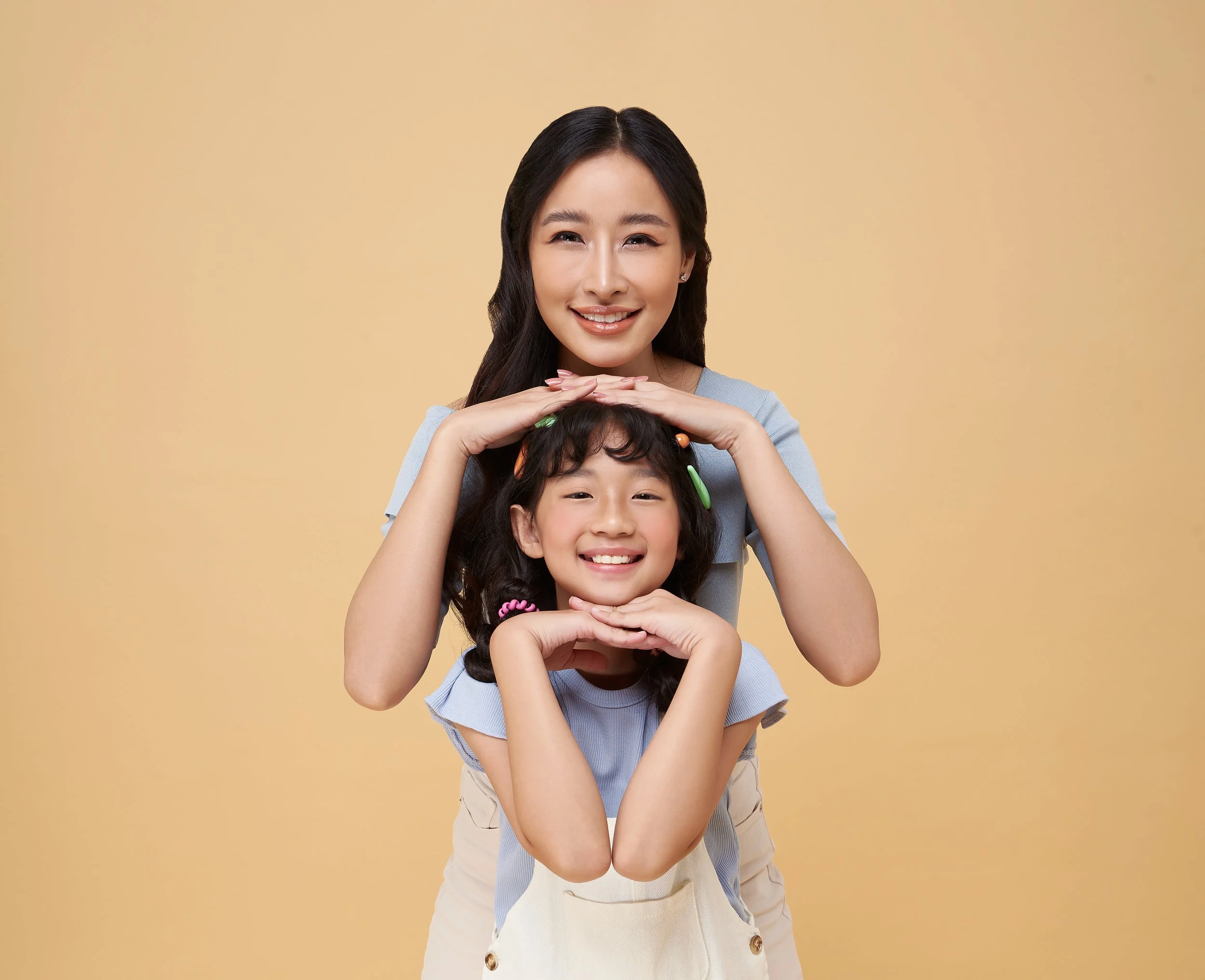 A woman and a girl smiling, with the woman standing behind the girl, resting her hands on the girl's head, against a beige background.