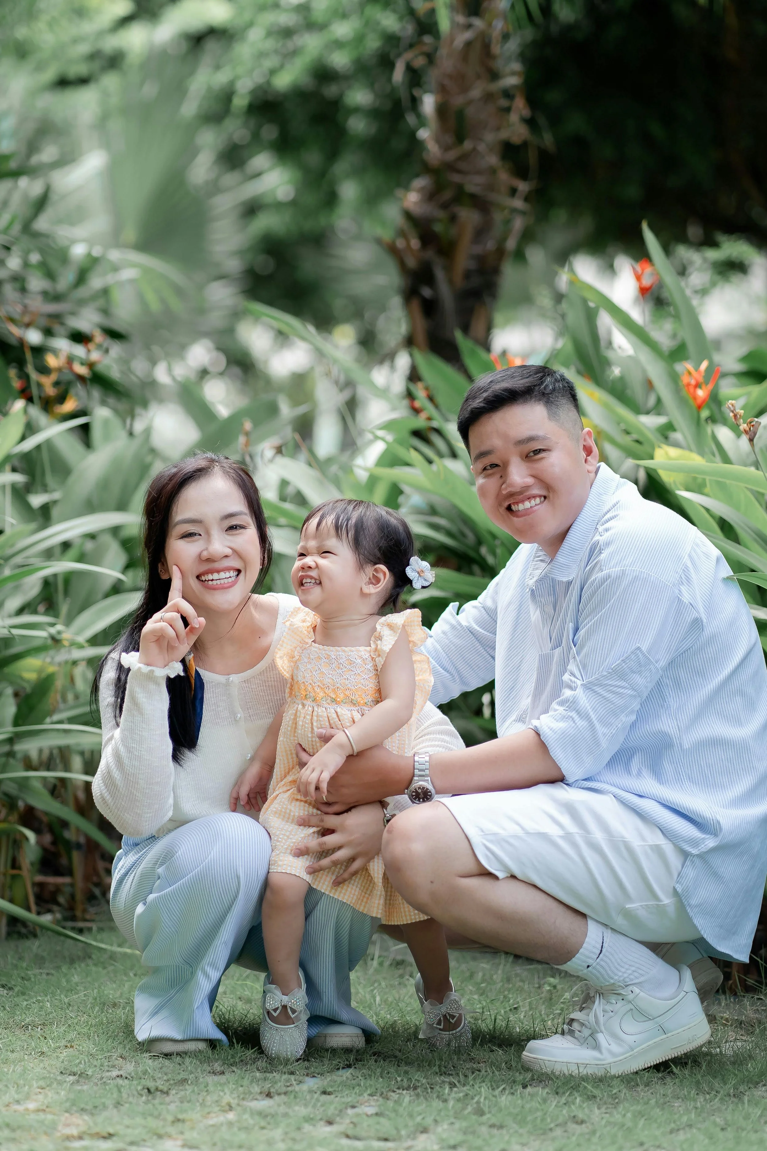 Family of three smiling and sitting outdoors among green plants, with a woman, man, and young girl in casual clothing.