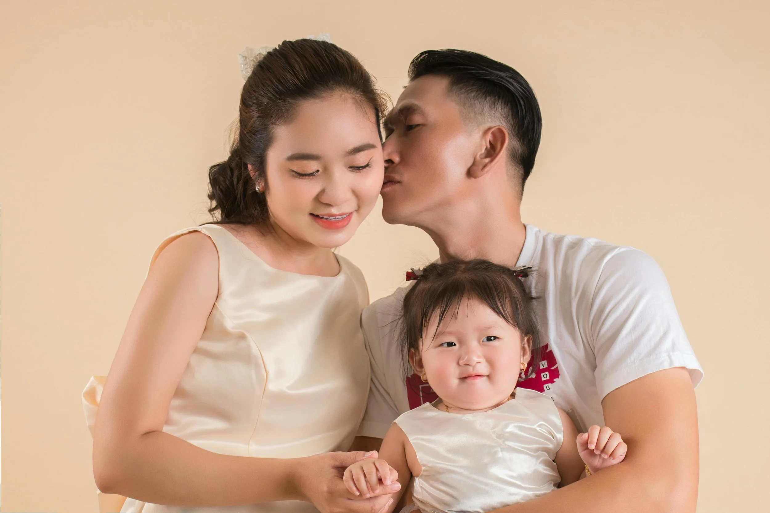 A family of three, with a woman, a man, and a young girl, poses together. The man is kissing the woman on the cheek, and the young girl is smiling at the camera while holding hands with the woman. The family appears happy and close.