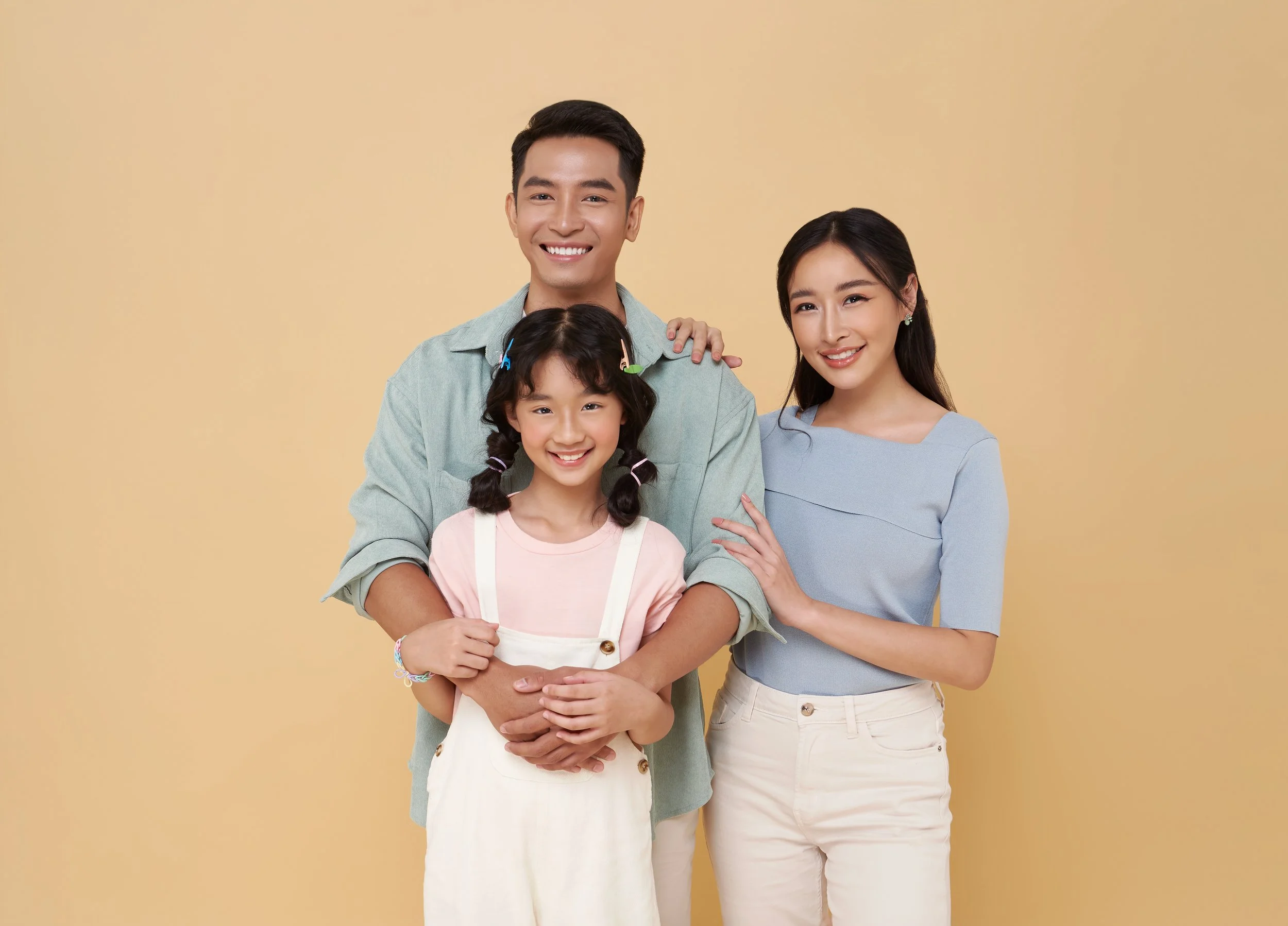 A happy family of three, a man, woman, and young girl, standing together against a plain beige background.