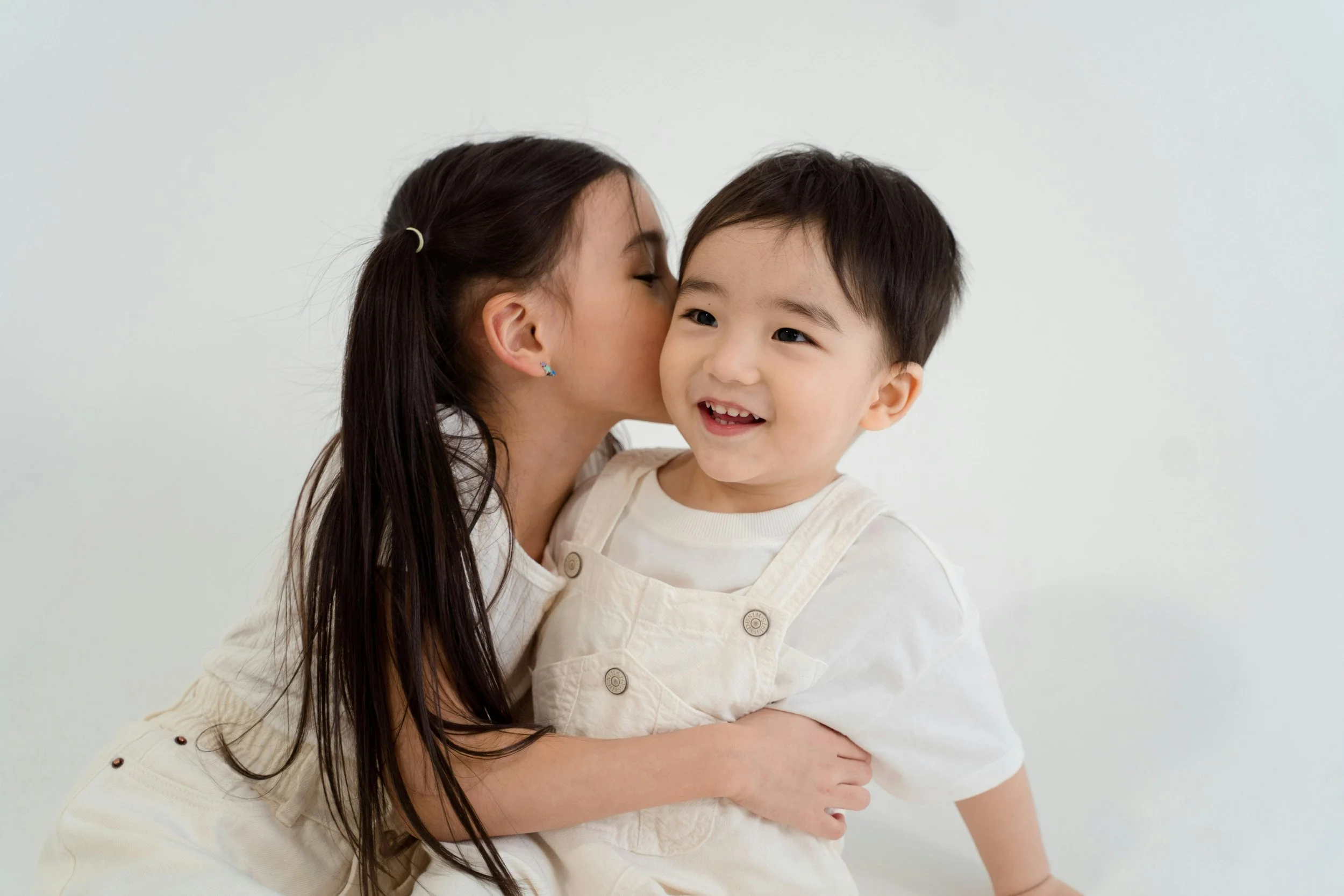 A young girl kissing a smiling young boy on the cheek while holding him, both dressed in light-colored clothing, against a plain white background.