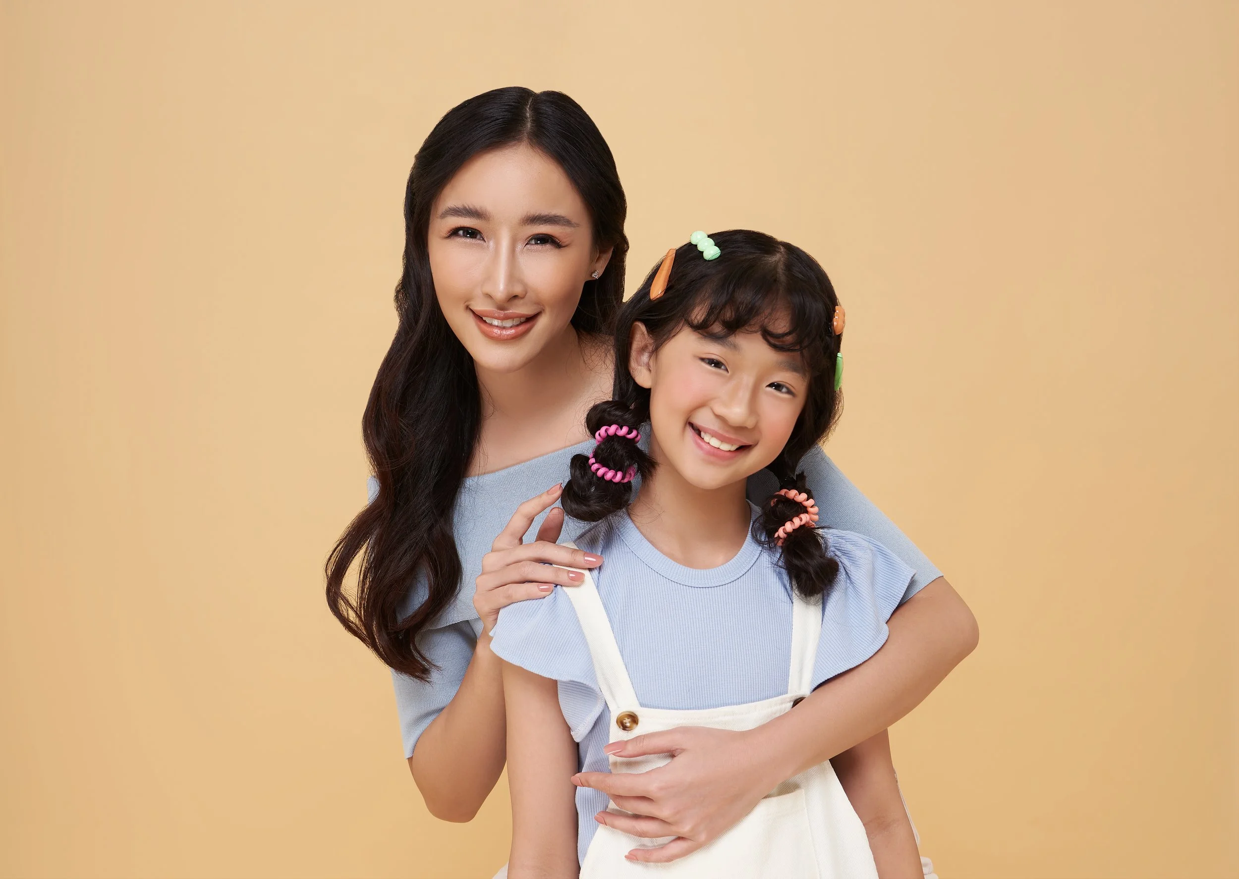 A young woman and girl smiling and hugging against a beige background, with the girl having colorful hair clips and pigtails.