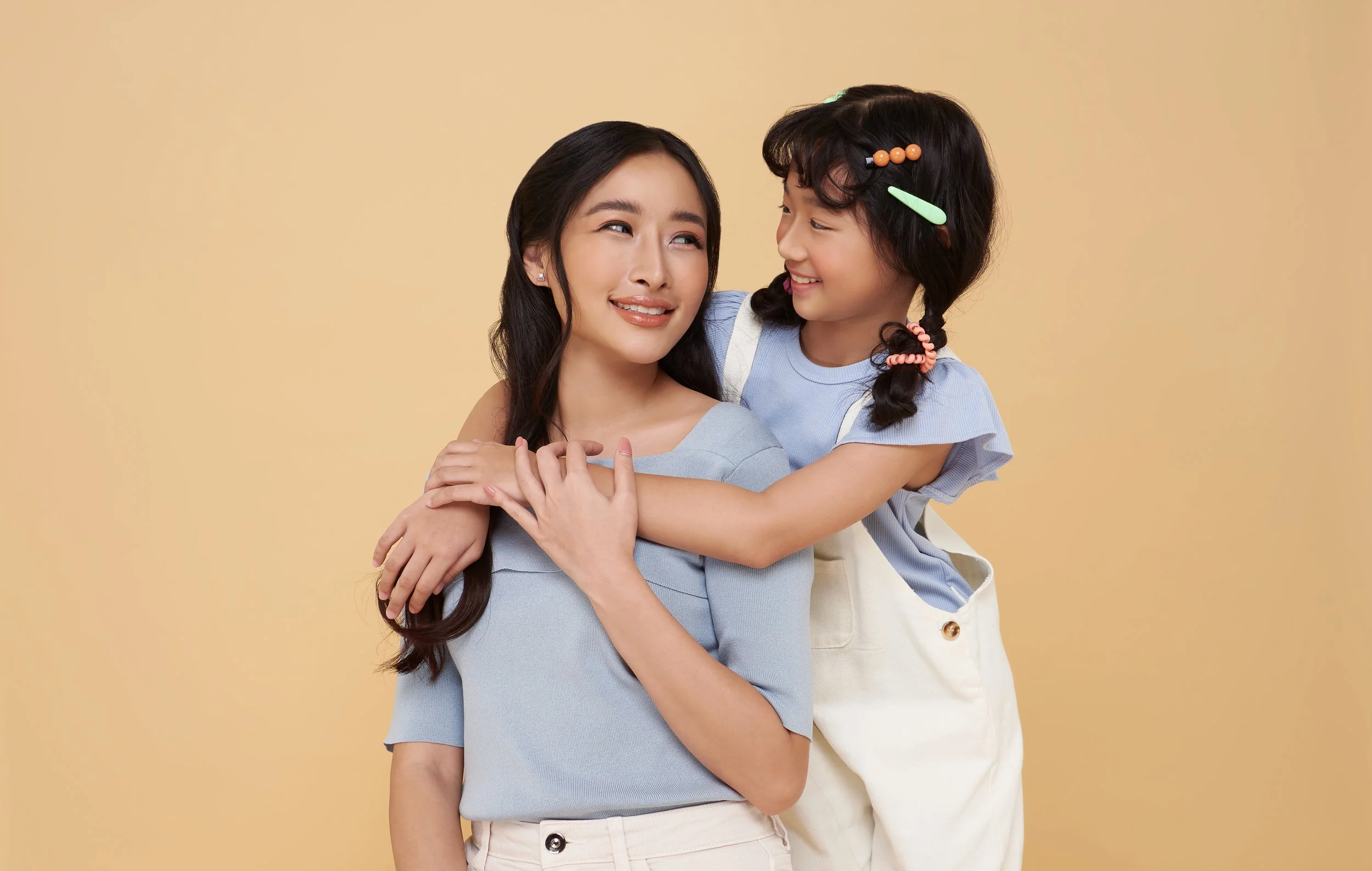A woman and a young girl sharing a joyful moment, with the girl hugging the woman's shoulders. The woman is smiling softly, and the girl is smiling cheerfully with colorful hair clips in her braided hair. They are against a plain, light beige backgro