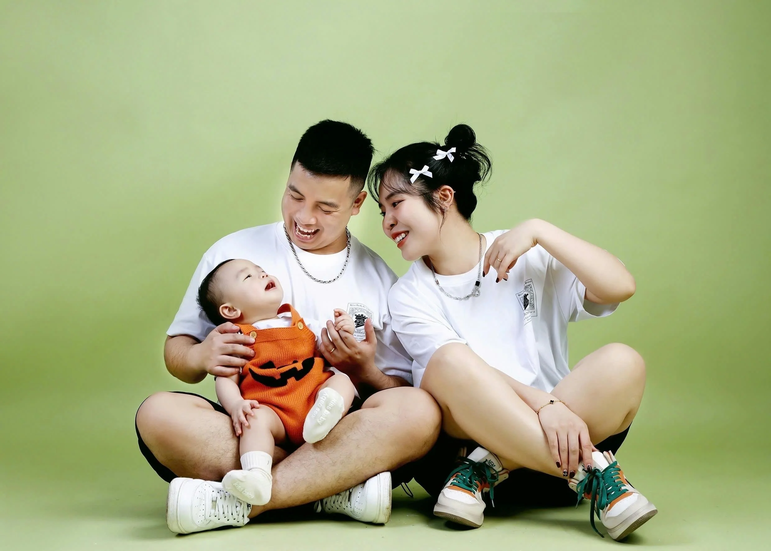 A happy family of three sitting on the floor against a light green background. The father and mother are smiling and looking at their baby, who is dressed in a Halloween-themed orange outfit with a pumpkin face. The mother has her hair in pigtails wi