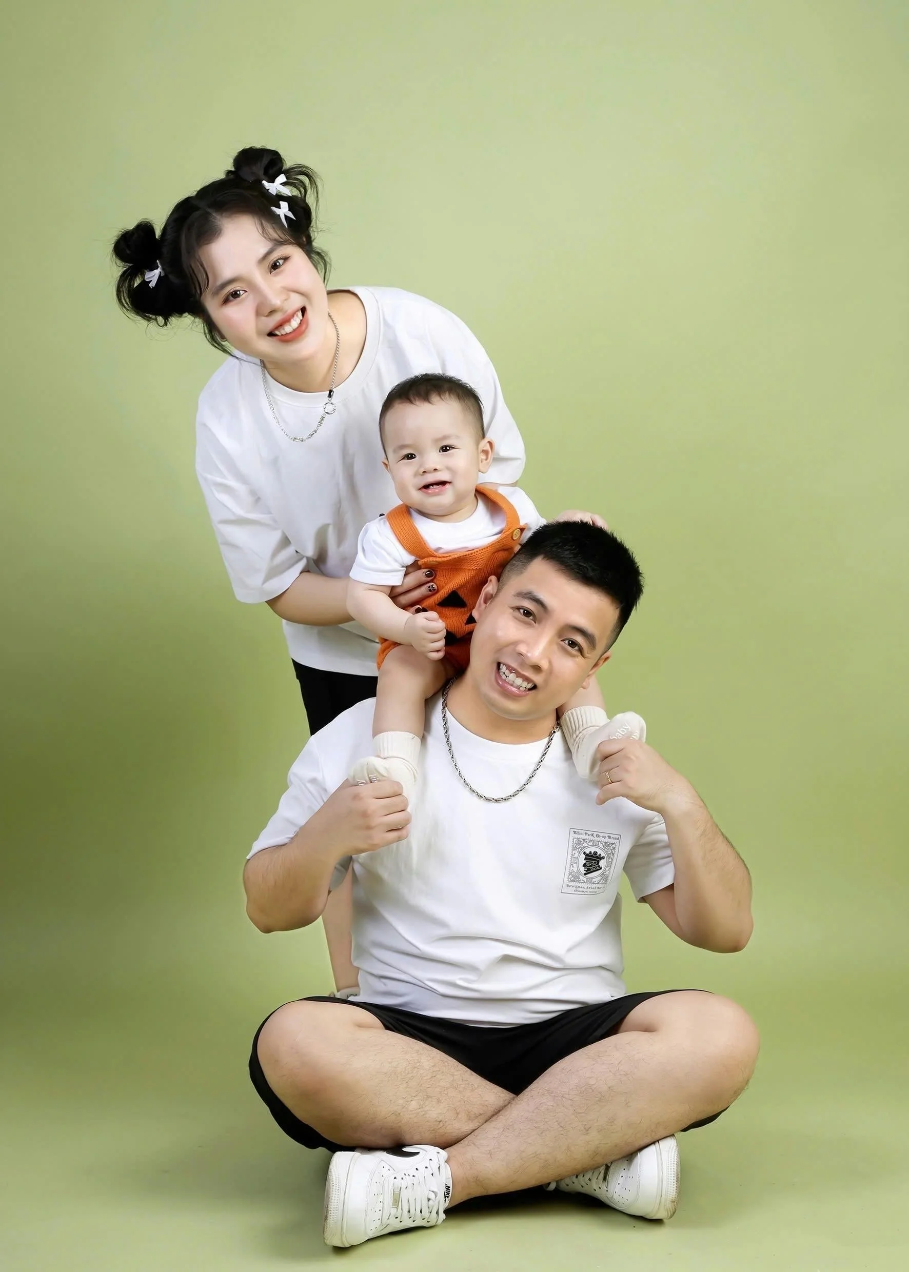 Family of three with a mother, father, and a young child sitting on the floor against a green background. The mother has black hair styled in playful pigtails with white ribbons, and the father has short black hair. The child, perched on the father’s