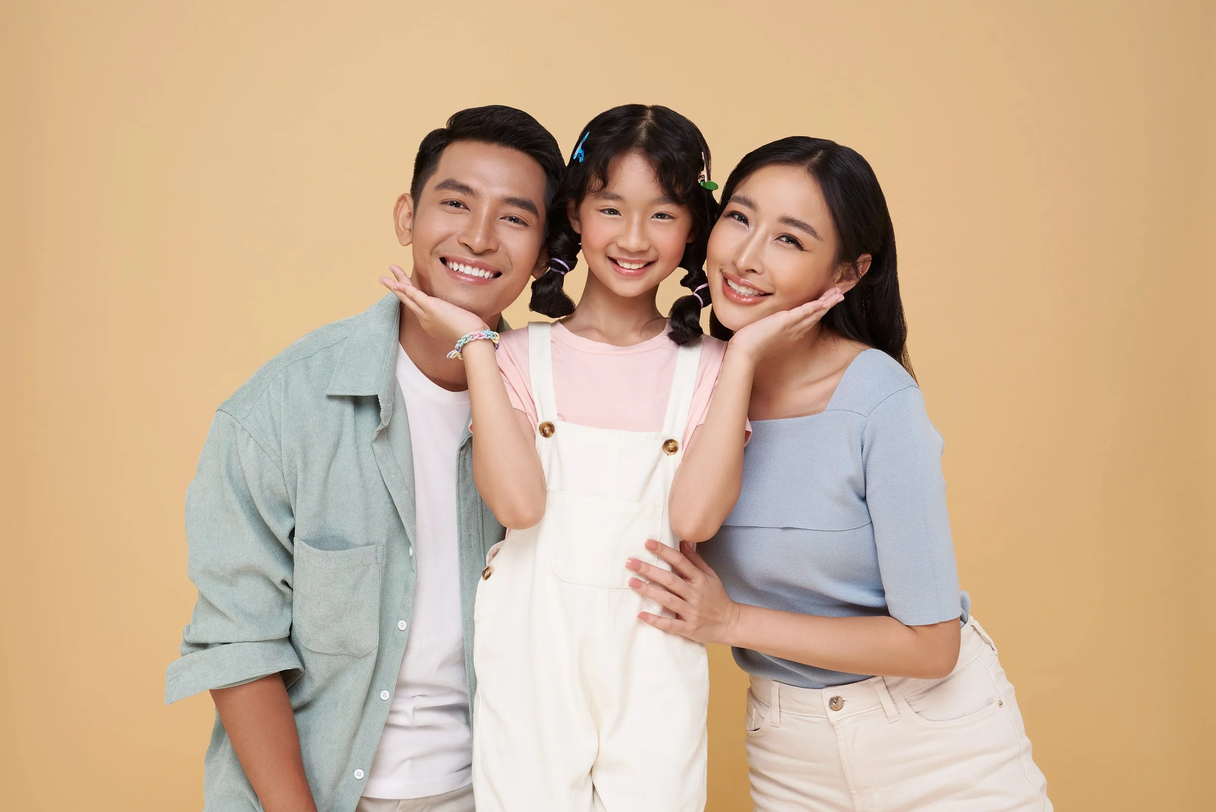 A smiling family of three, a man, a woman, and a girl, posing together against a yellow background, with the girl in the center smiling and touching her cheeks, flanked by the parents.