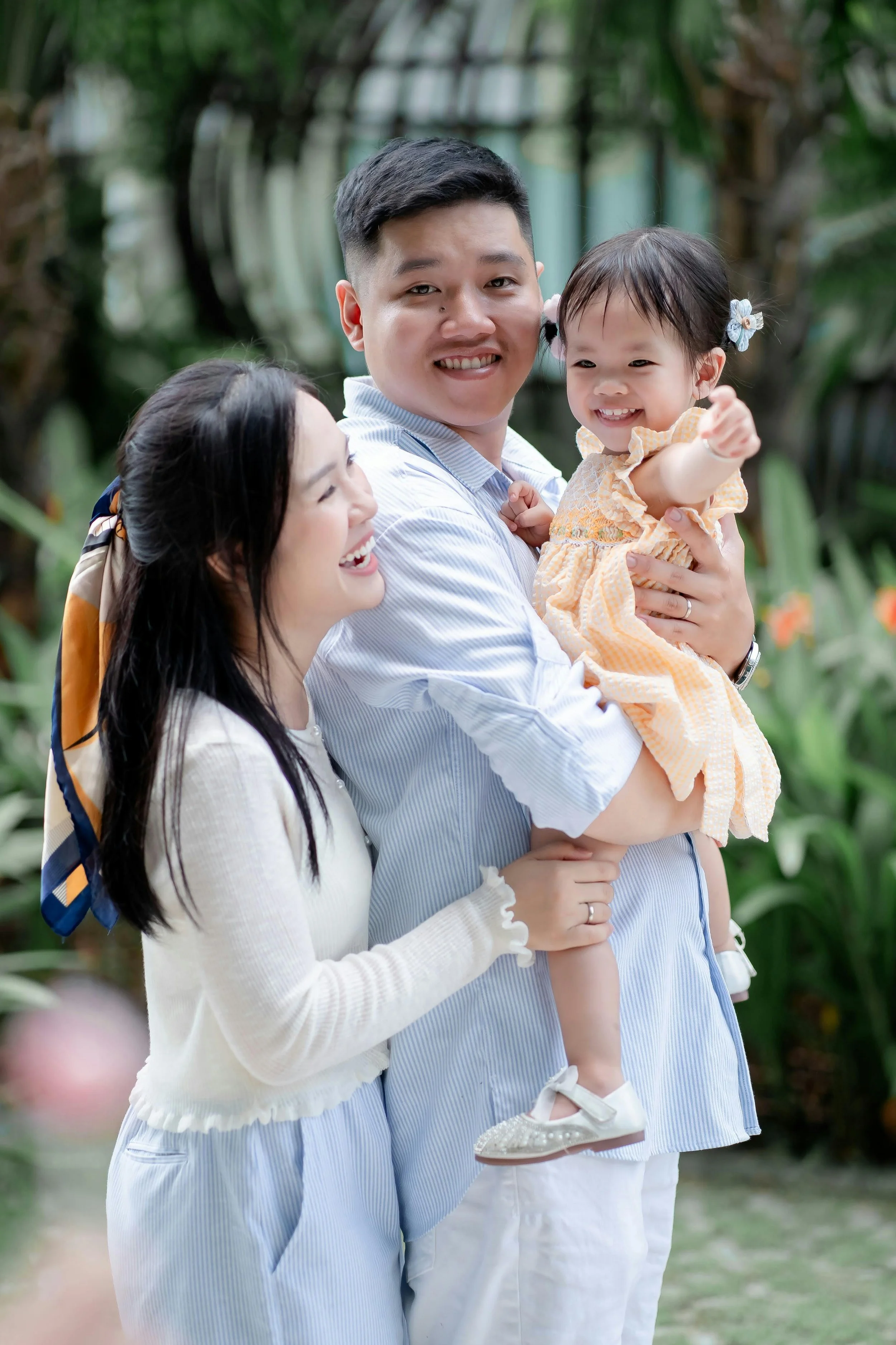 A happy family of three, with a man holding a young girl, and a woman smiling at them outdoors in a garden.