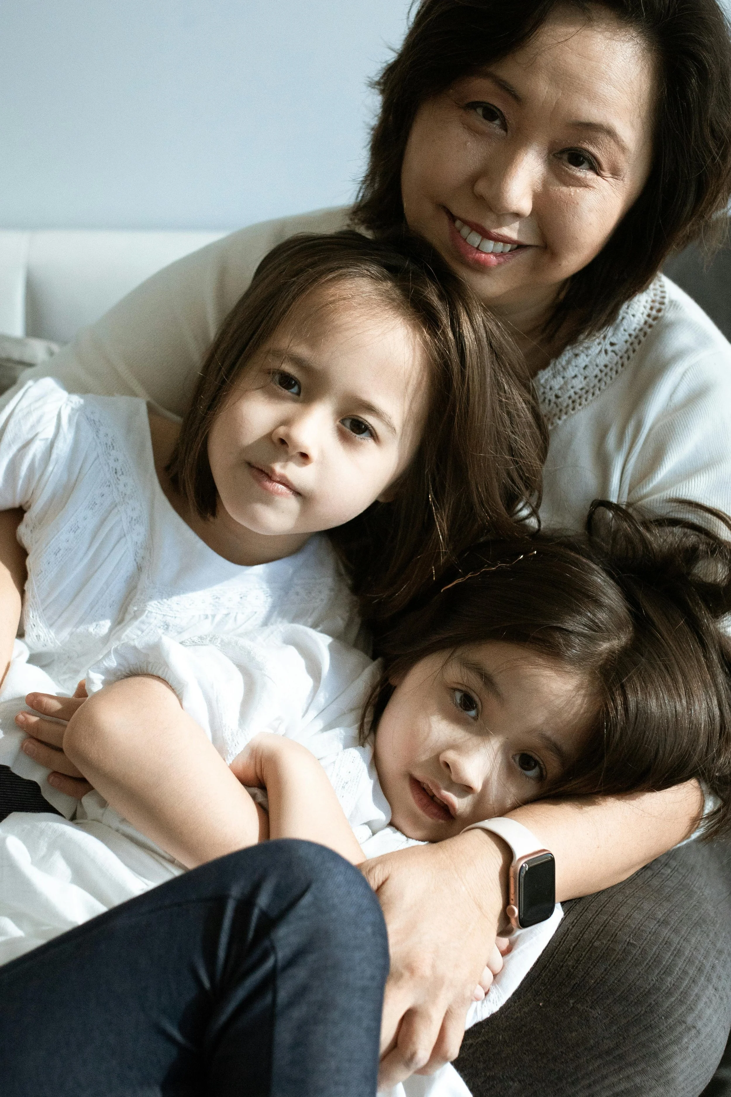 A woman with short dark hair smiling and hugging two young girls, all wearing white tops, in a cozy indoor setting.