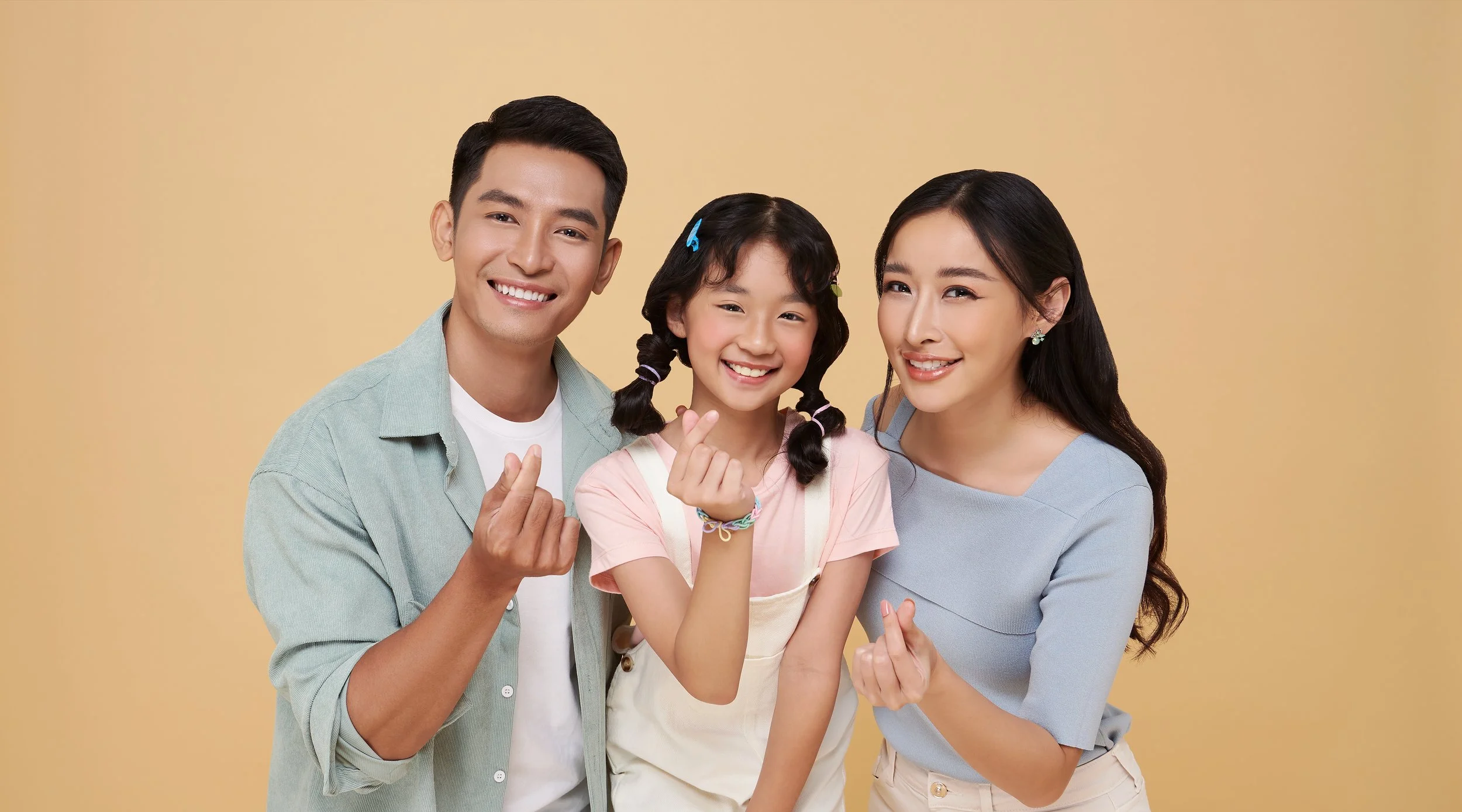 A smiling Asian family of three making heart hand gestures against a beige background