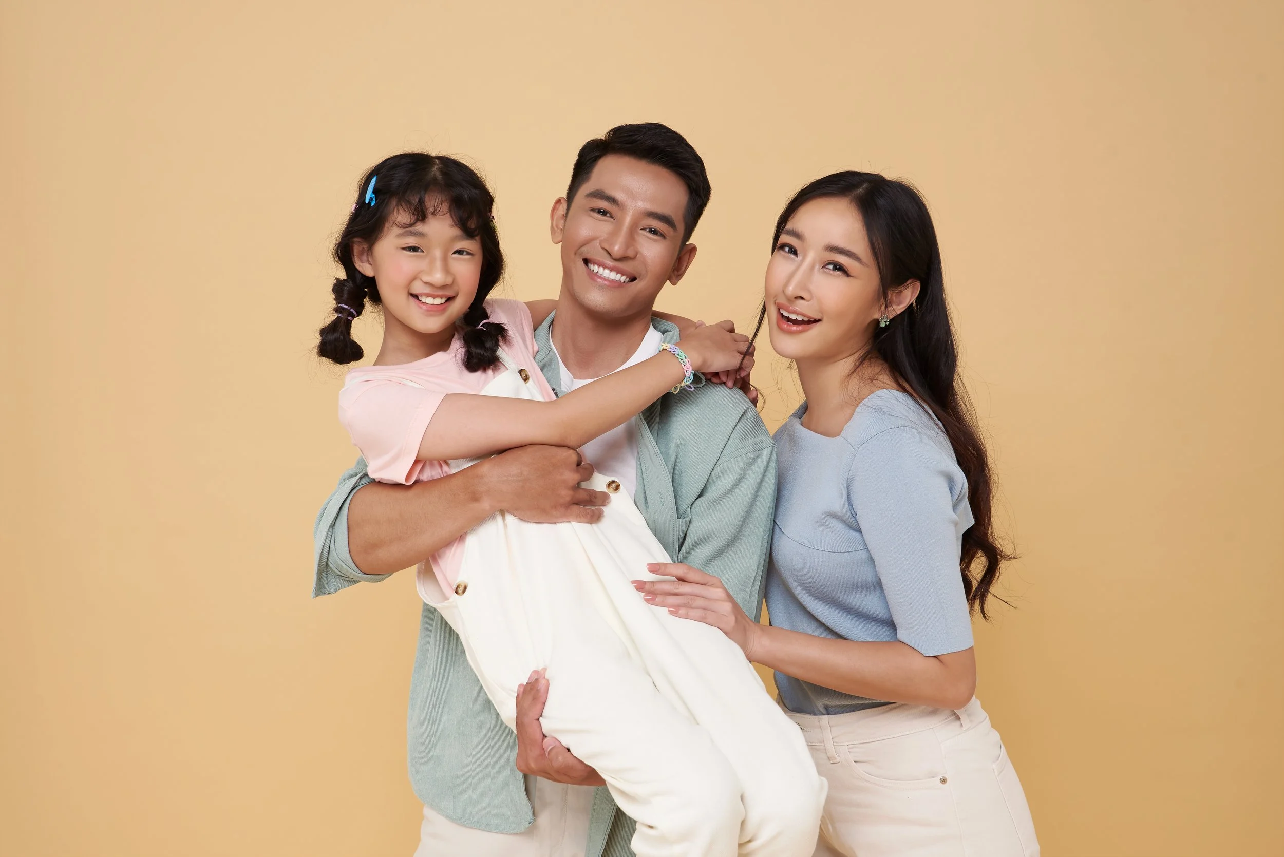 A happy family of three posing against a yellow background, with a man holding a young girl and a woman standing beside them.
