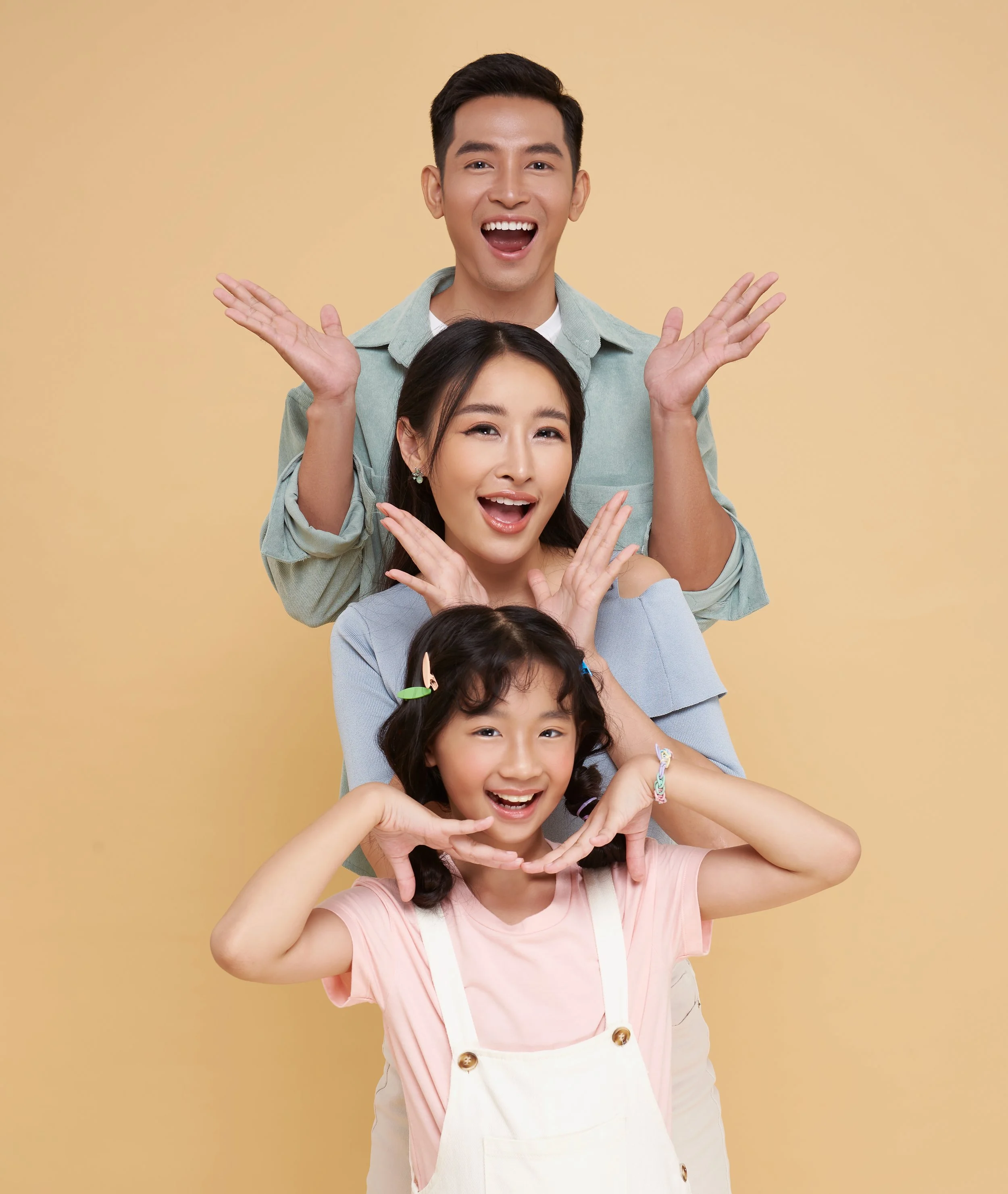 A joyful family of four stacked in a vertical line against a beige background, smiling and making playful gestures with their hands.