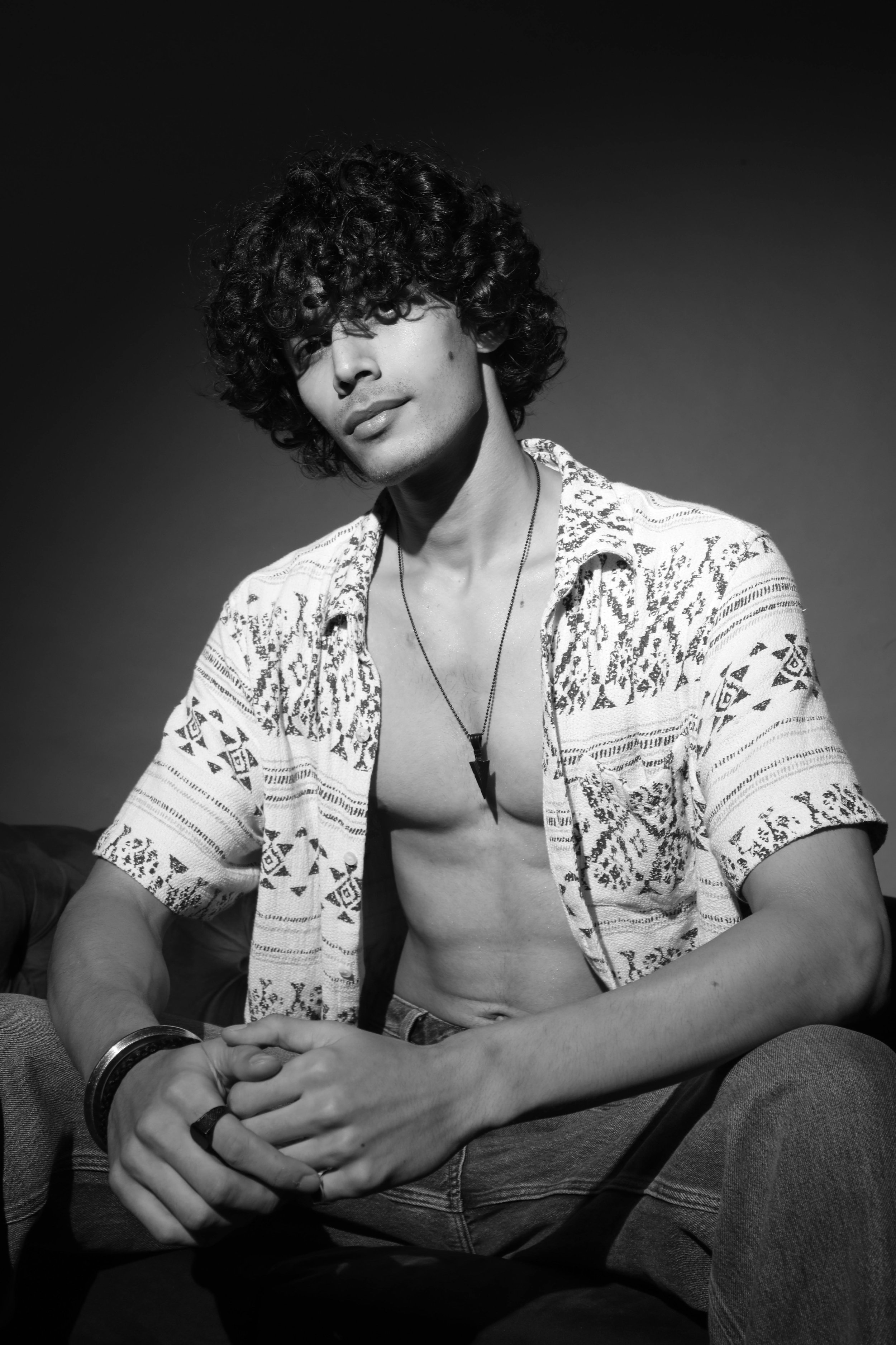 A young man with curly hair, wearing a patterned short-sleeve shirt and jeans, sitting with his hands clasped in his lap, looking at the camera in a black and white studio portrait.