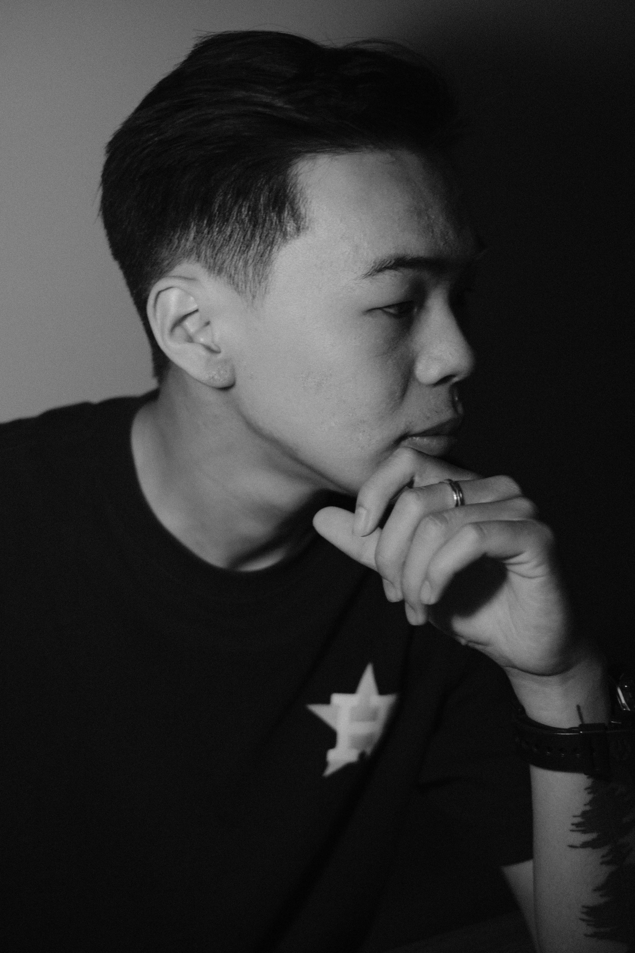 Black and white photo of a young man with styled hair, wearing a watch and rings, resting his chin on his hand, with tattooed arm visible.