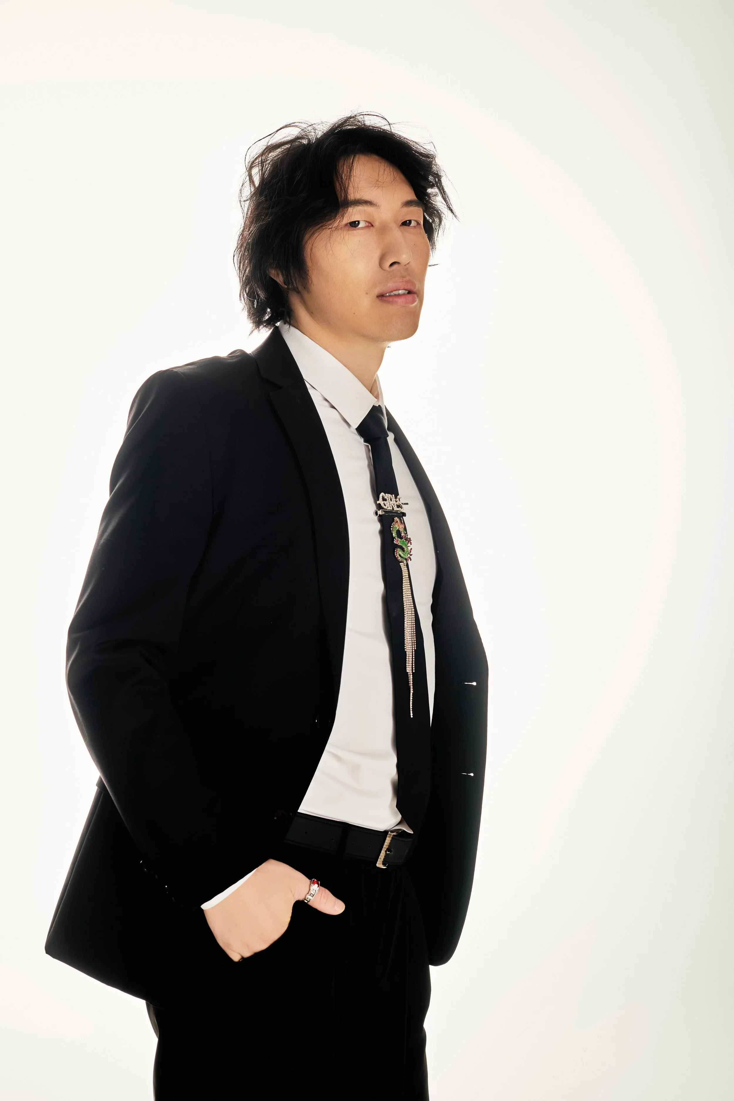 A man in a black suit with a white shirt and black tie, standing with one hand in his pocket, against a plain white background.