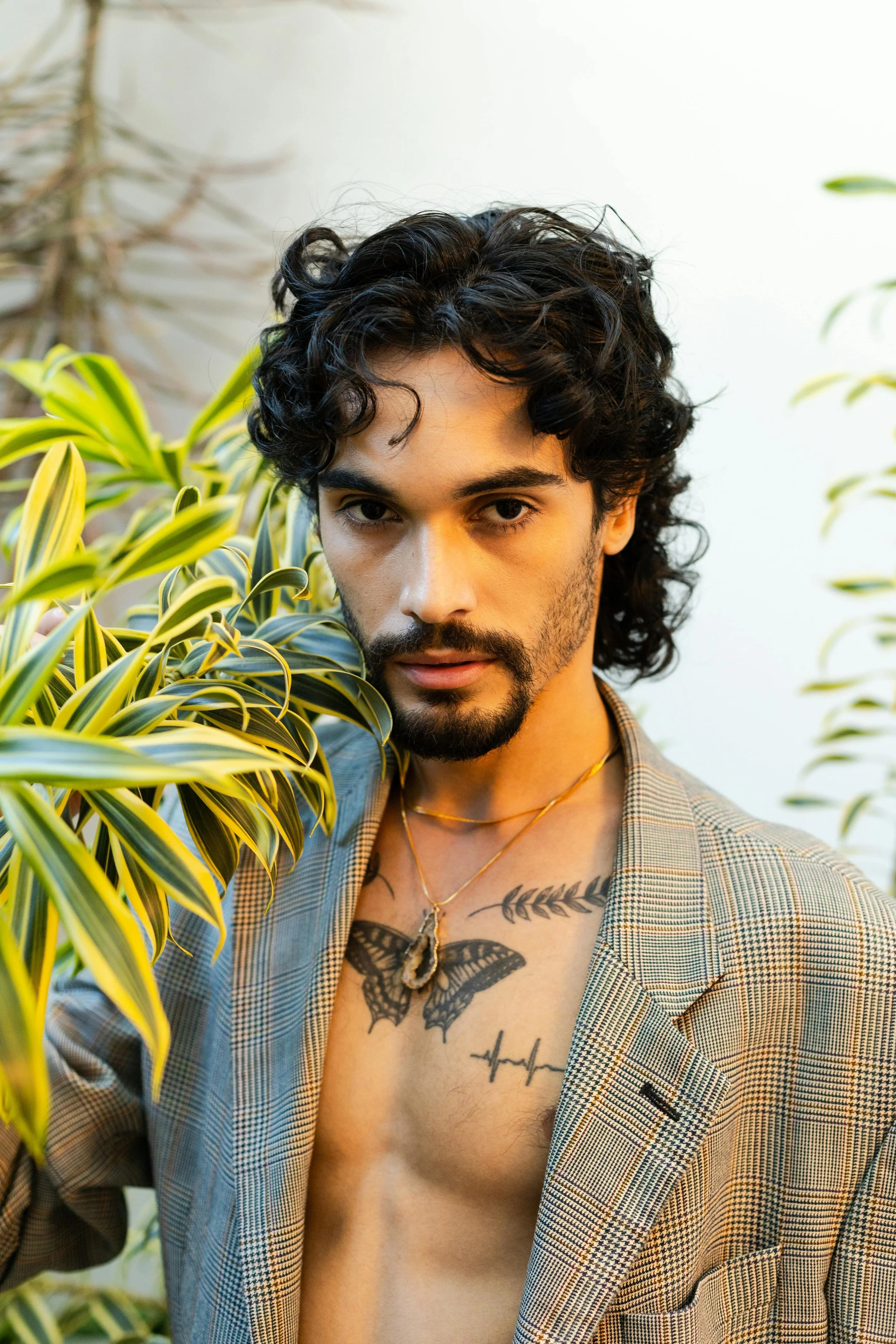 A man with dark curly hair and a beard, wearing a checkered blazer and gold necklace, poses shirtless next to a green leafy plant, showing tattoos on his chest, including a butterfly, a leaf branch, and an electrocardiogram line.