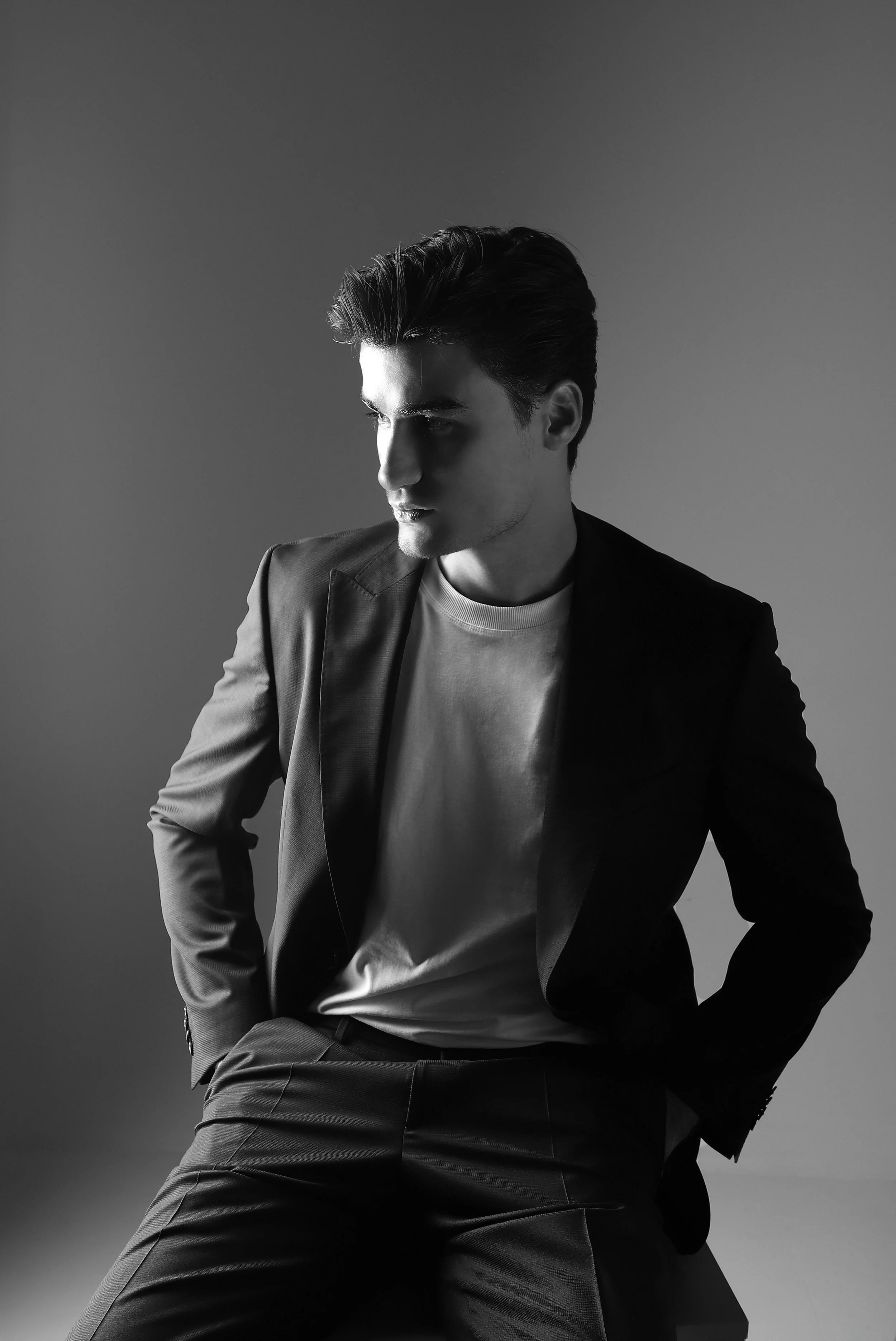 Black and white photo of a young man with dark hair, dressed in a blazer and white t-shirt, looking down thoughtfully, with hands in his pockets, against a plain background.