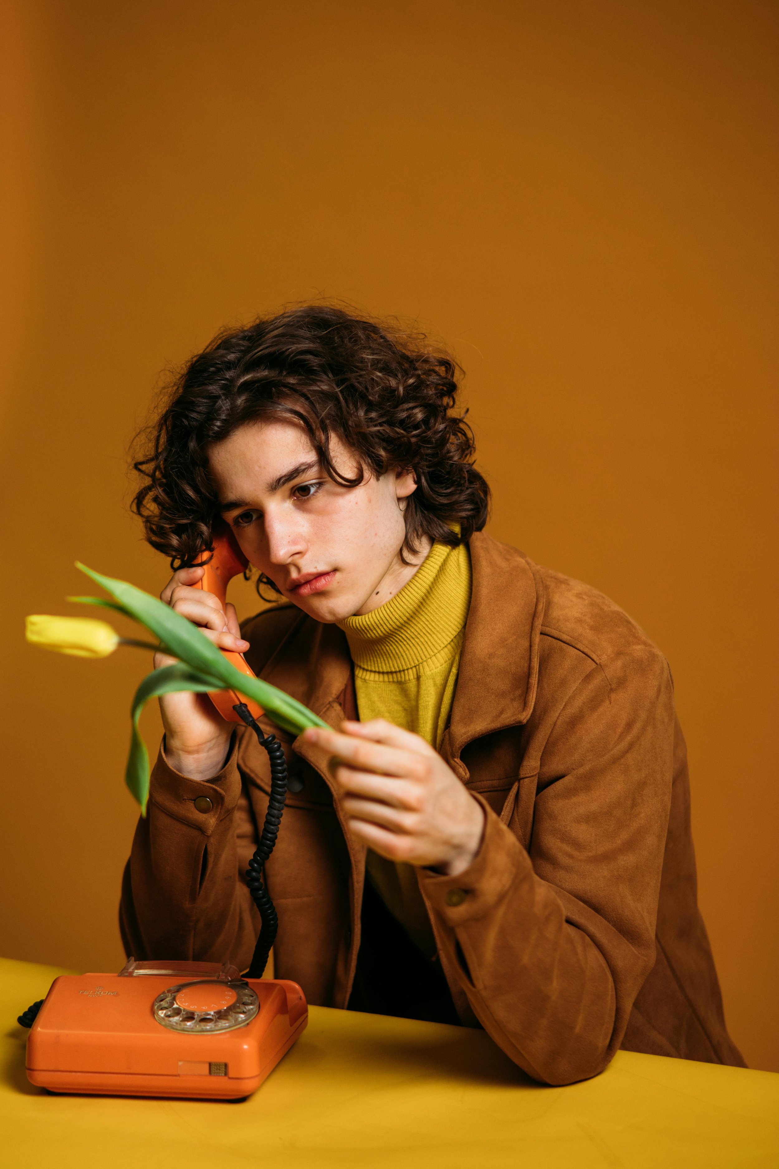 A young person with curly hair, wearing a brown jacket and yellow turtleneck, holding a yellow tulip and talking on an orange rotary phone against a yellow background.