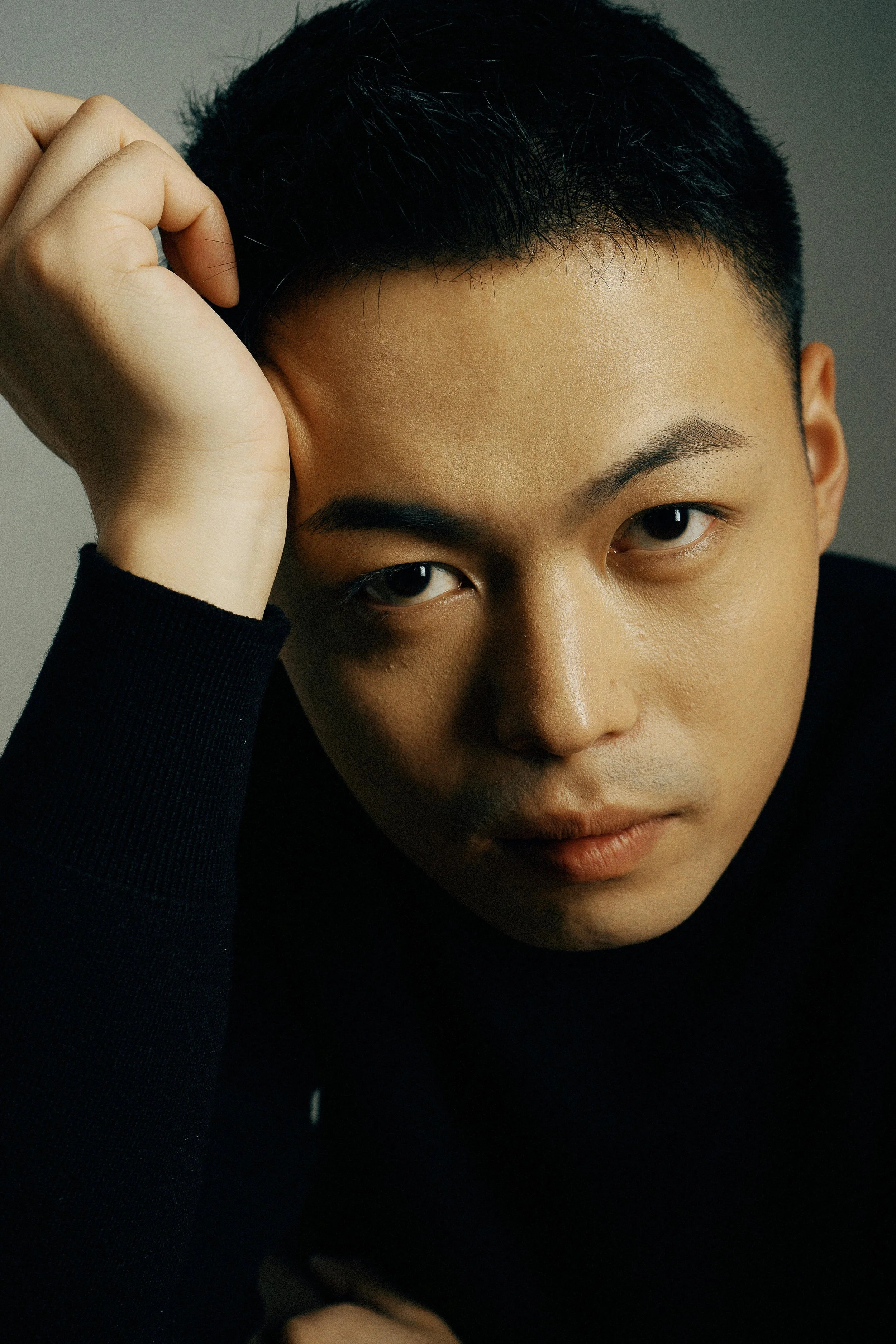 A close-up portrait of a young man with short black hair, wearing a black shirt, looking intently at the camera with his head slightly tilted and resting his hand on his forehead.