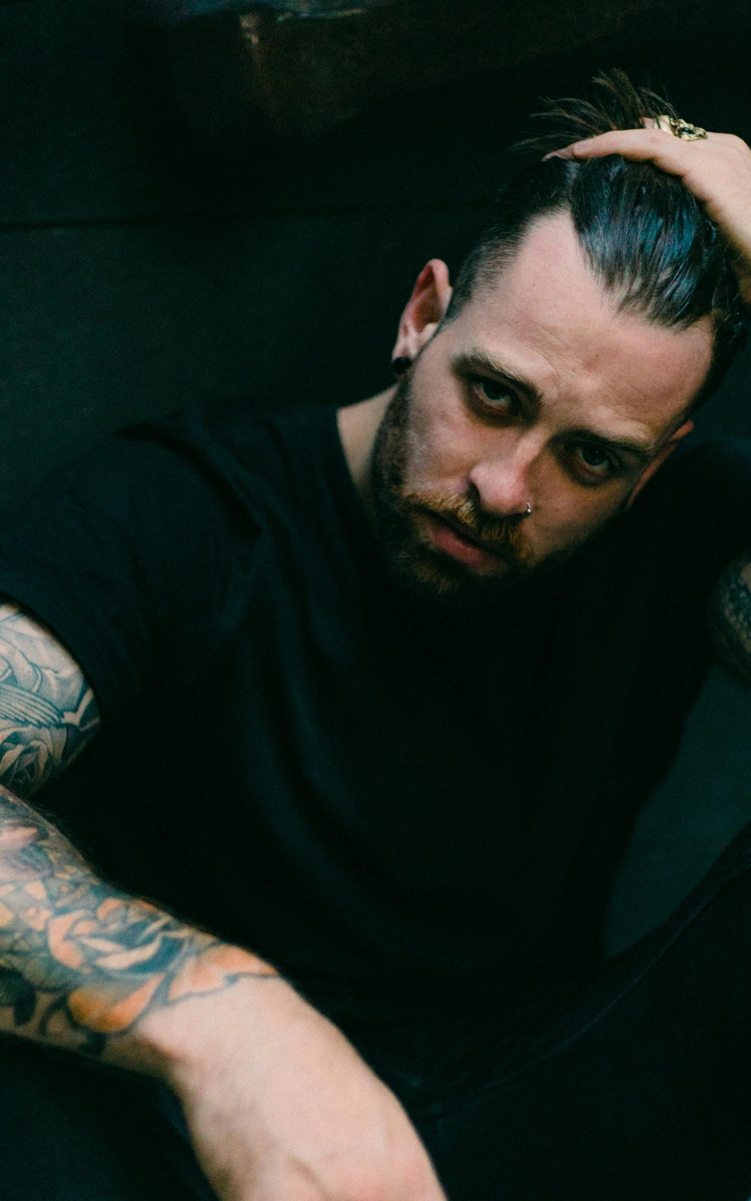 Portrait of a man with tattoos, dark hair, and facial piercing, lying on a dark surface, with one hand on his head and wearing a black shirt.