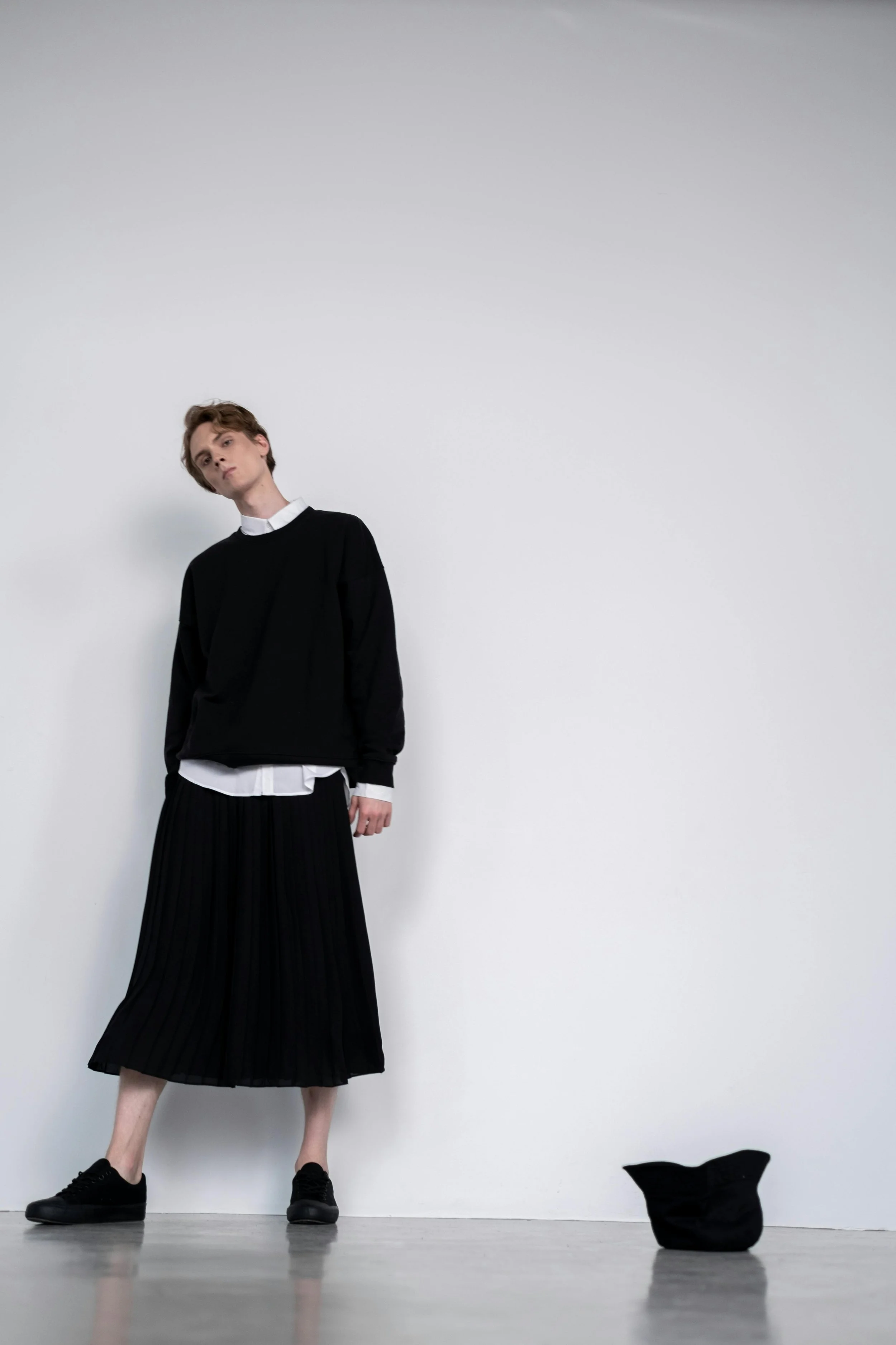 A person with short light brown hair and pale skin stands against a plain white wall, dressed in a black sweater layered over a white shirt, with a black pleated midi skirt and black shoes. A black hat lies on the ground nearby.
