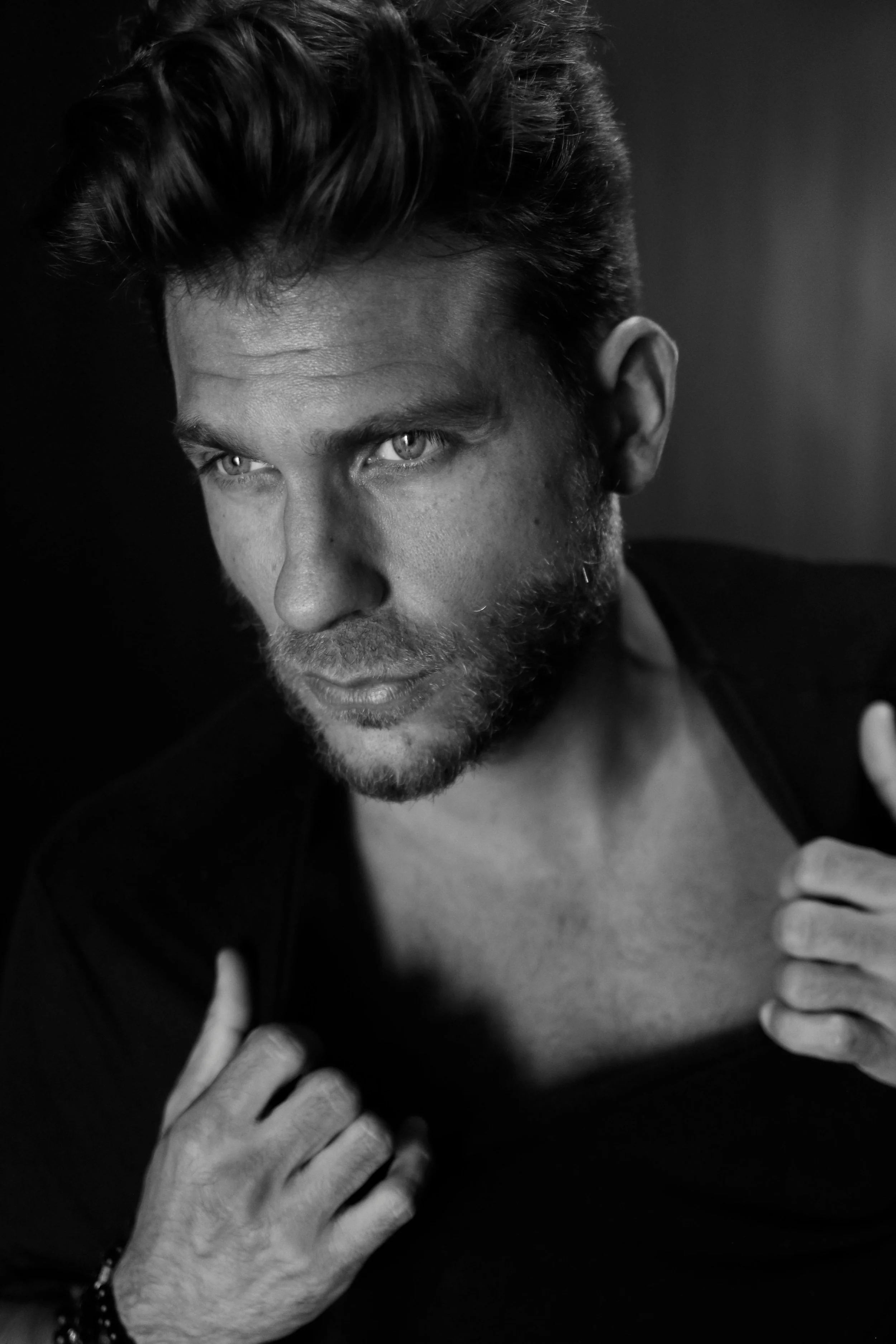 Black and white portrait of a man with styled hair and beard, looking intently to the side, with one hand gripping his shirt collar.
