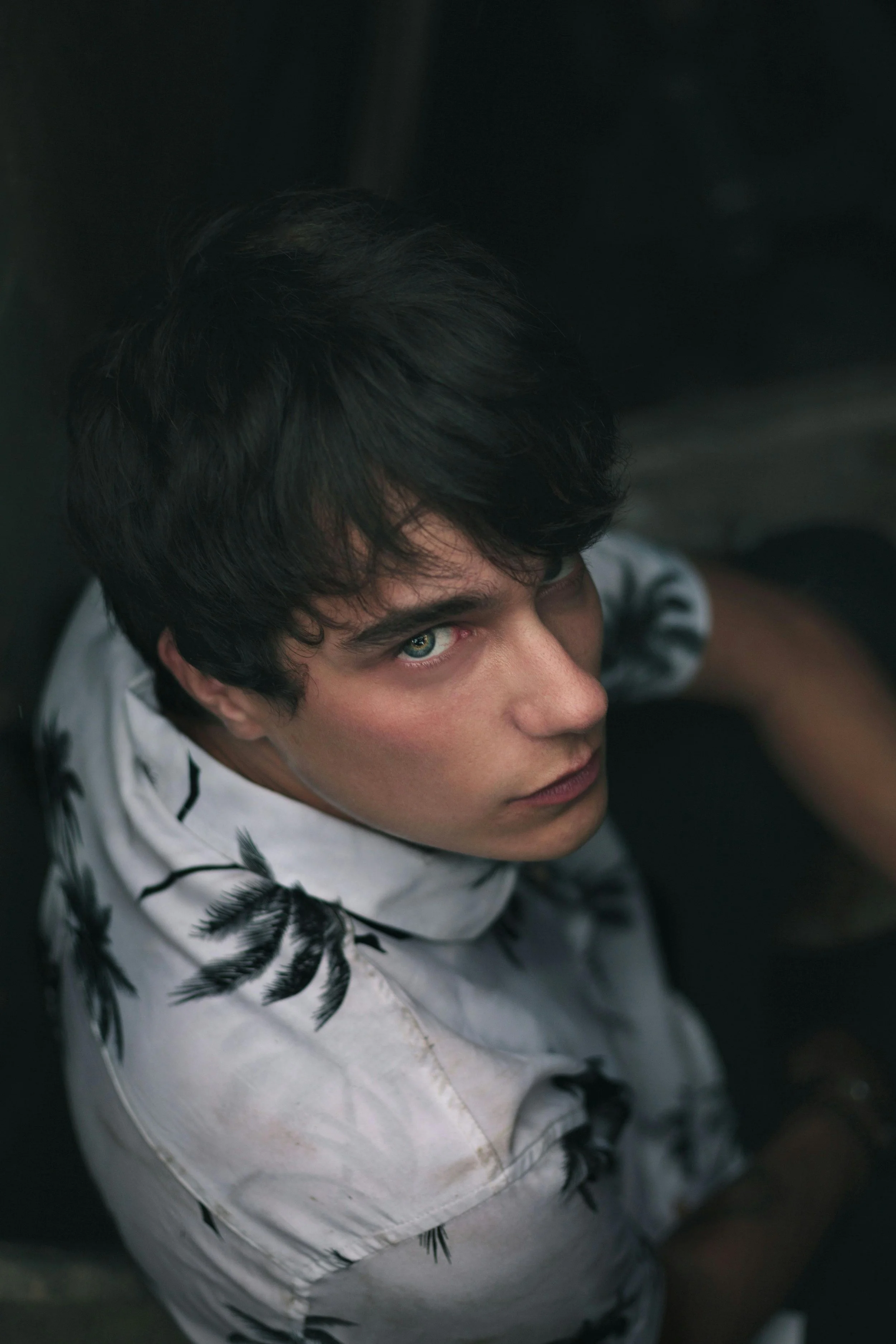 A young man with dark hair and blue eyes looking up at the camera, wearing a white shirt with a black leaf pattern, sitting in a dimly lit space.