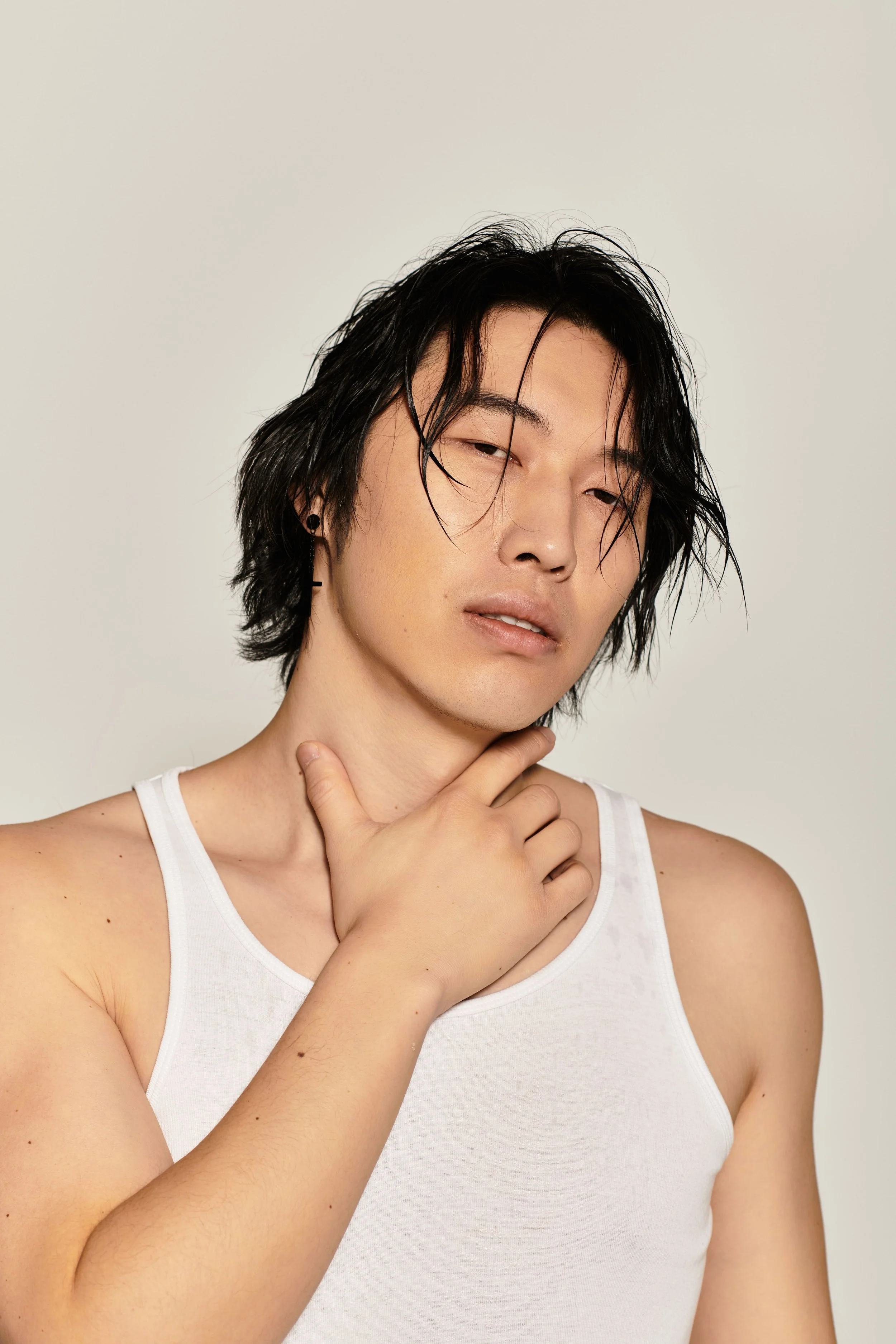 A person with wet, black, shoulder-length hair, wearing a white tank top, posing with one hand on their neck against a plain light background.