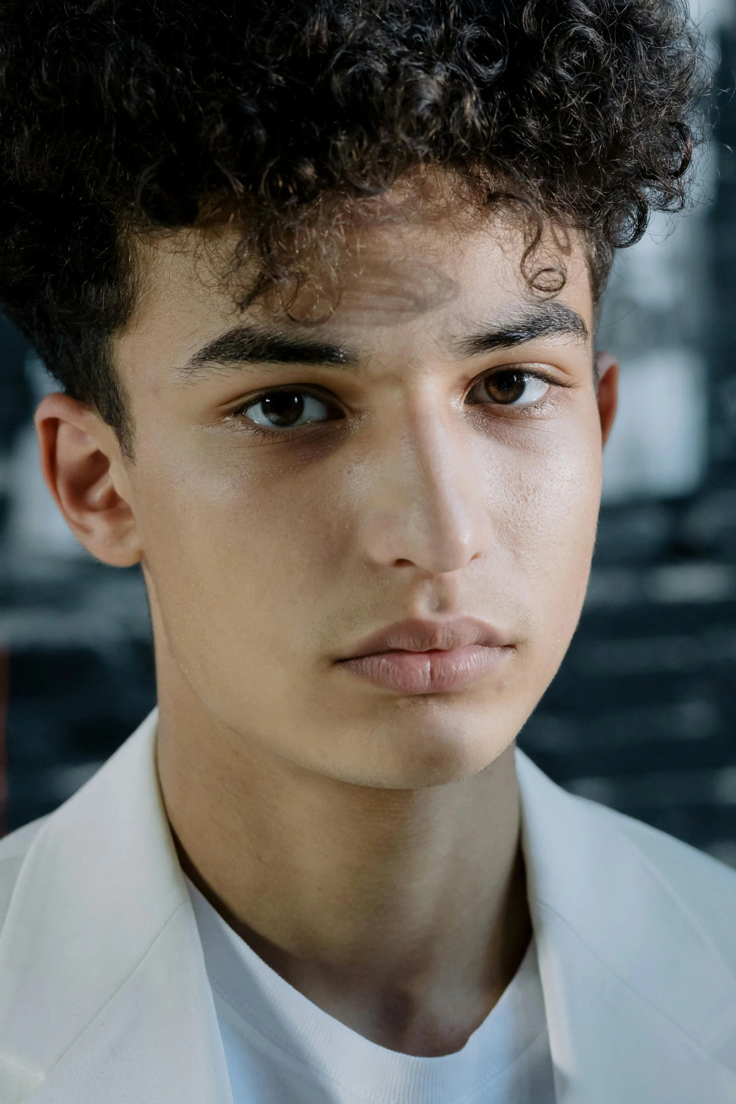 Close-up portrait of a young person with curly dark hair, brown eyes, and light skin, wearing a white shirt.
