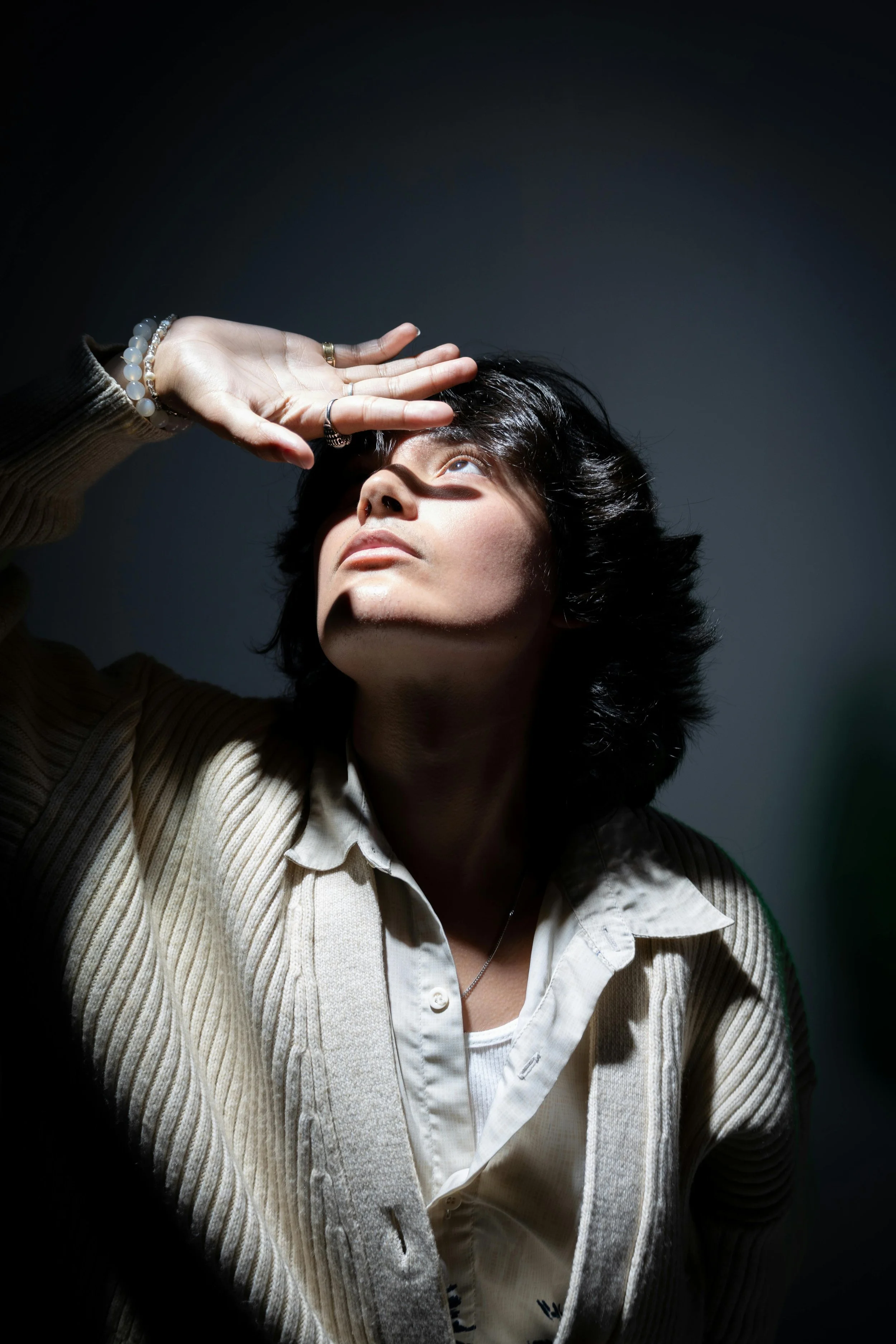 A woman with dark hair looking upward, shielding her eyes from bright light with her hand.