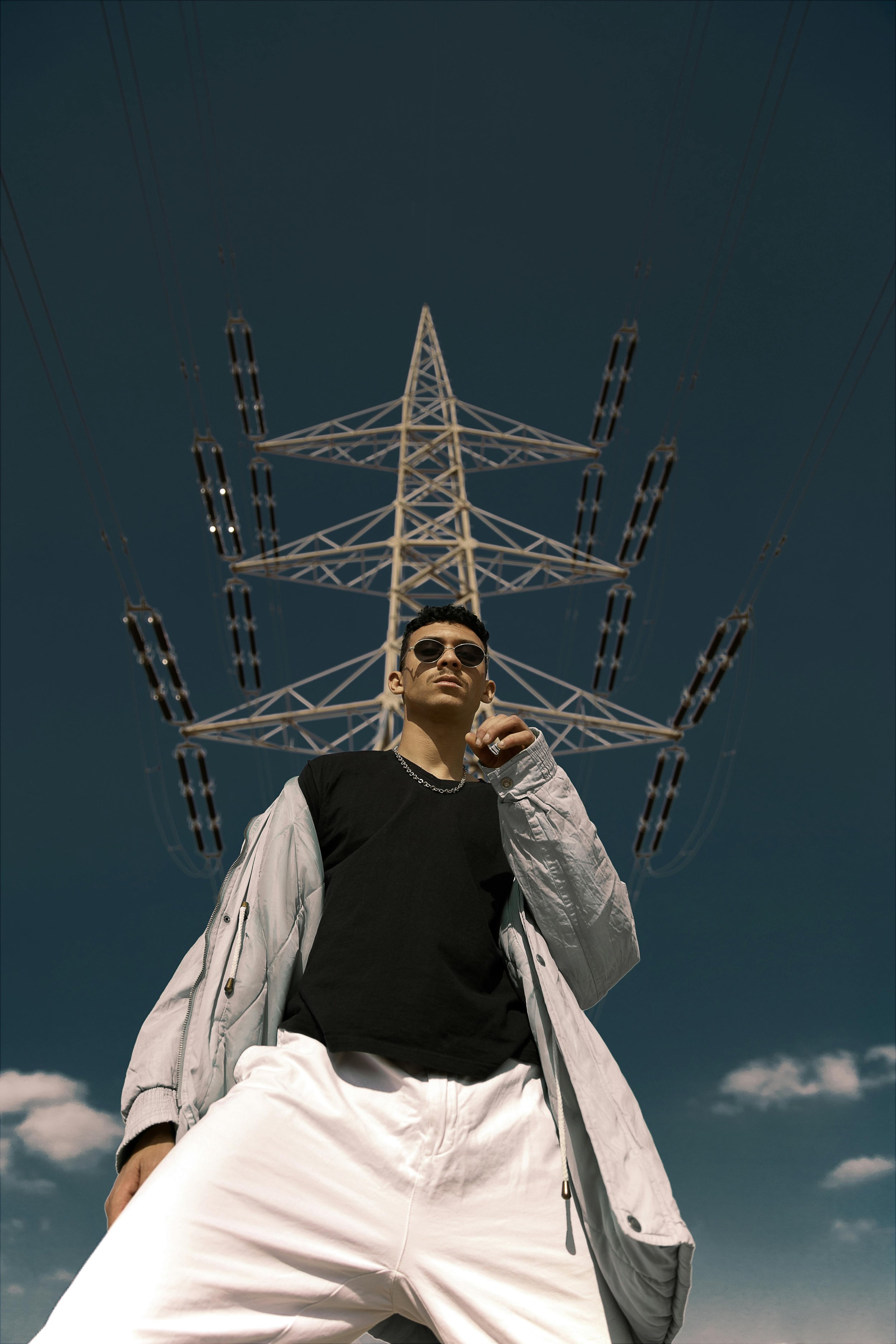 A young man wearing sunglasses, a black t-shirt, and a light-colored jacket over his shoulders stands outdoors with a large electrical transmission tower behind him against a blue sky with some clouds.