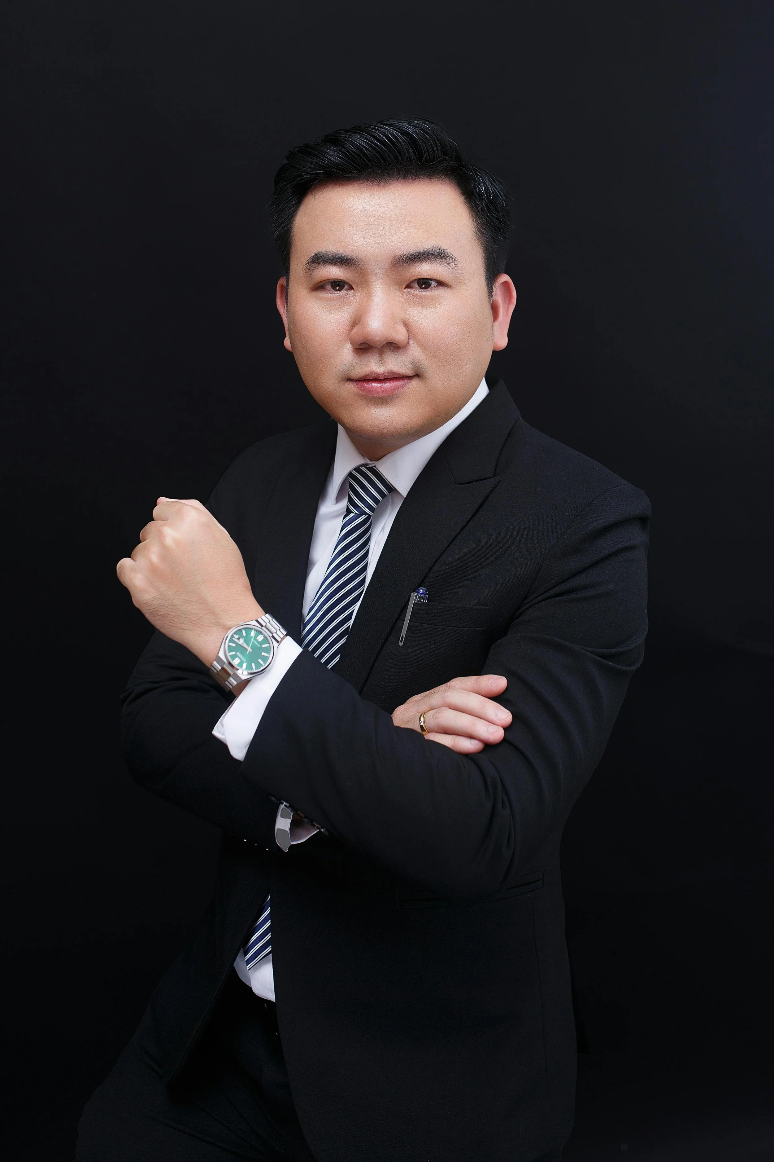 Professional portrait of a young man in a black suit, white shirt, striped tie, and wristwatch, posing against a black background.