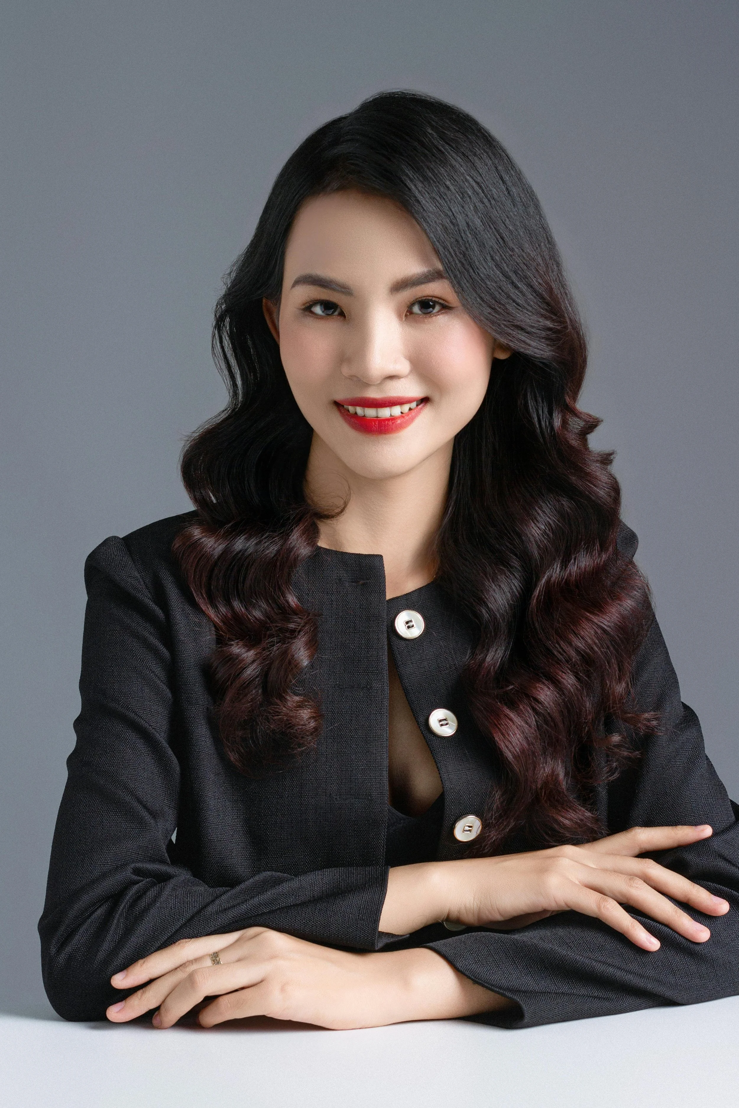 Portrait of a young Asian woman with wavy black hair, red lipstick, wearing a black blazer and smiling.