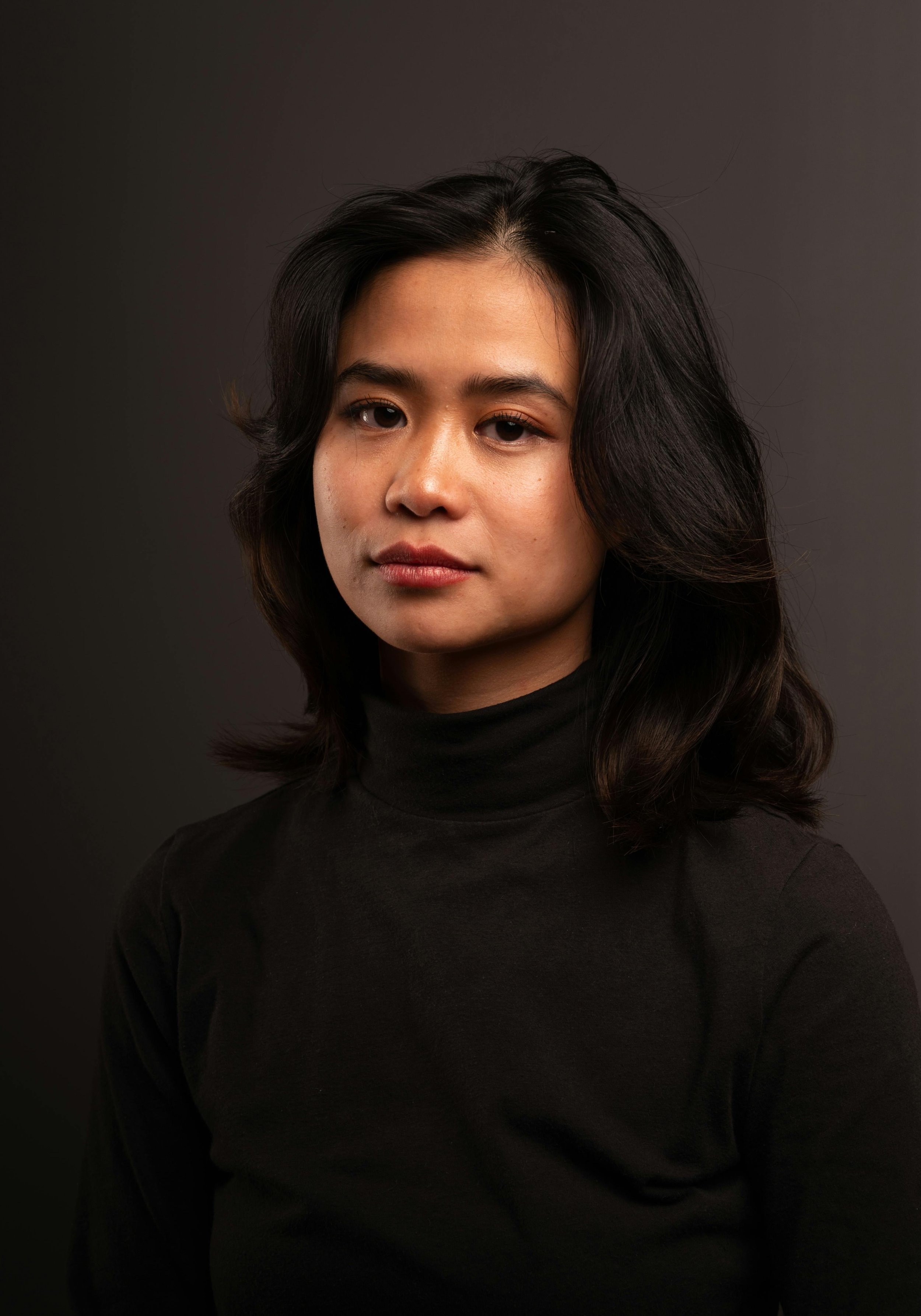 A woman with dark, wavy hair wearing a black turtleneck, looking at the camera with a neutral expression against a dark background.