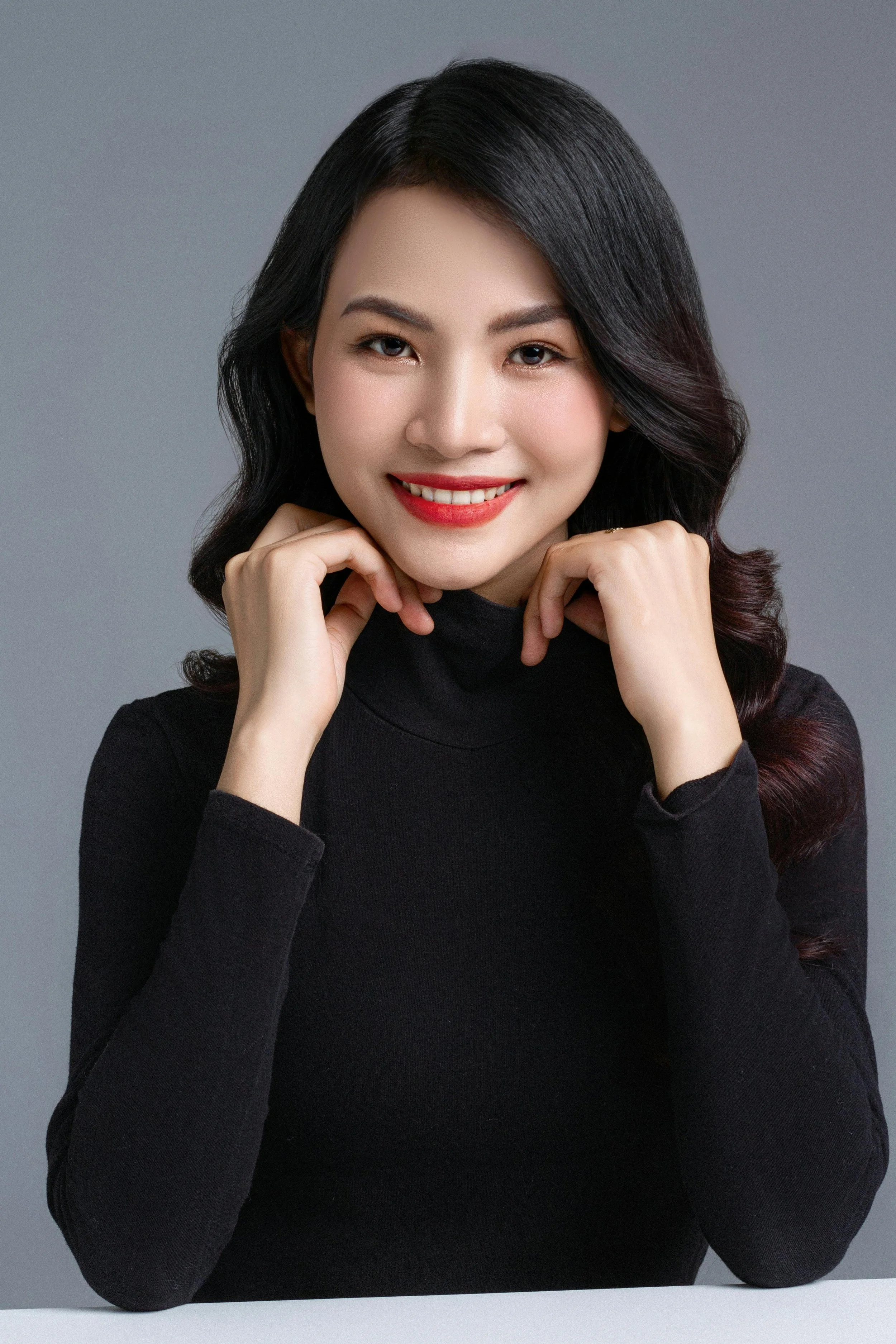A young woman with shoulder-length dark hair, wearing a black turtleneck, smiling and resting her chin on her hands against a plain gray background.