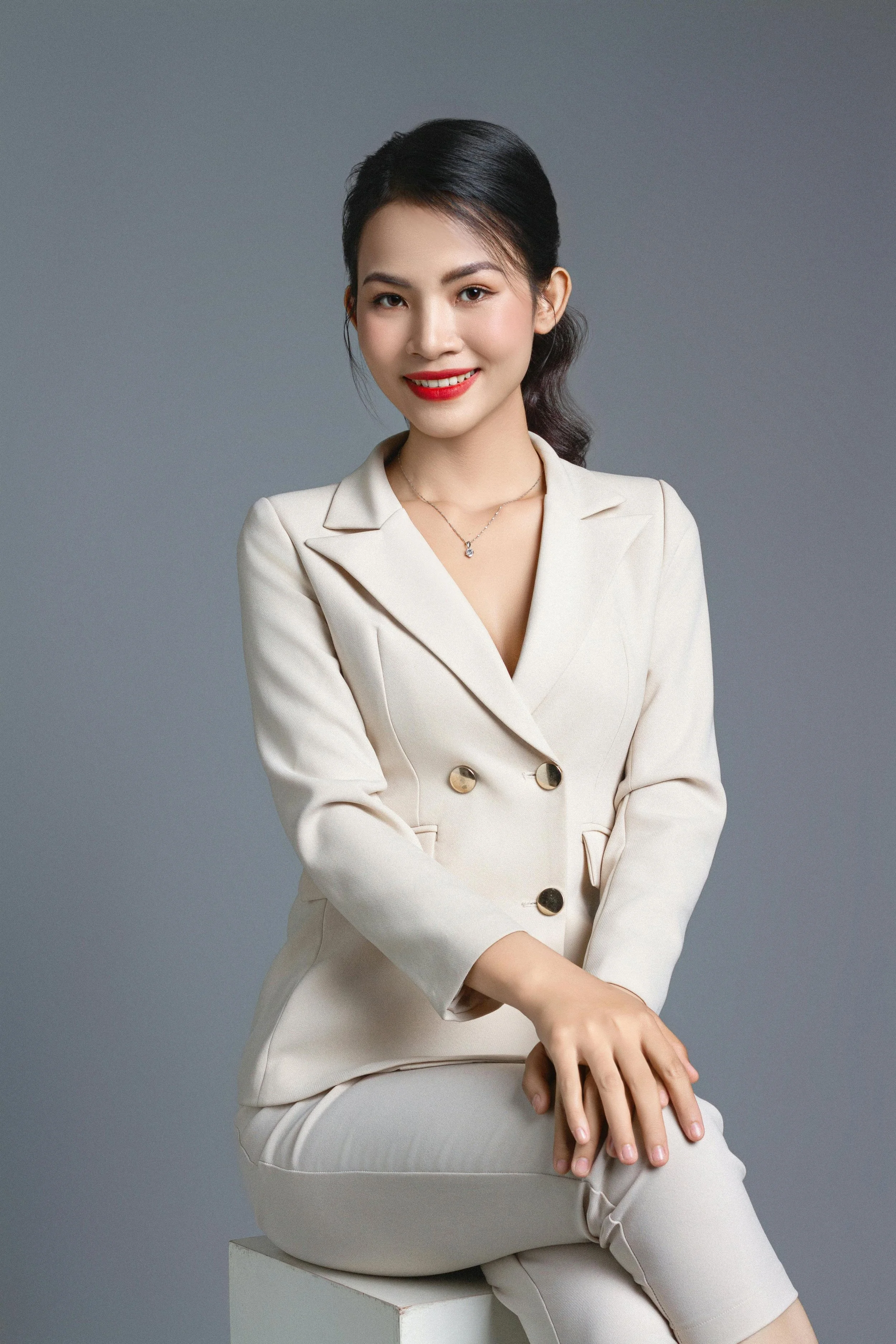 A young woman with dark hair styled in a ponytail, wearing a cream-colored blazer and pants, smiling with red lipstick, sitting on a white block against a gray background.