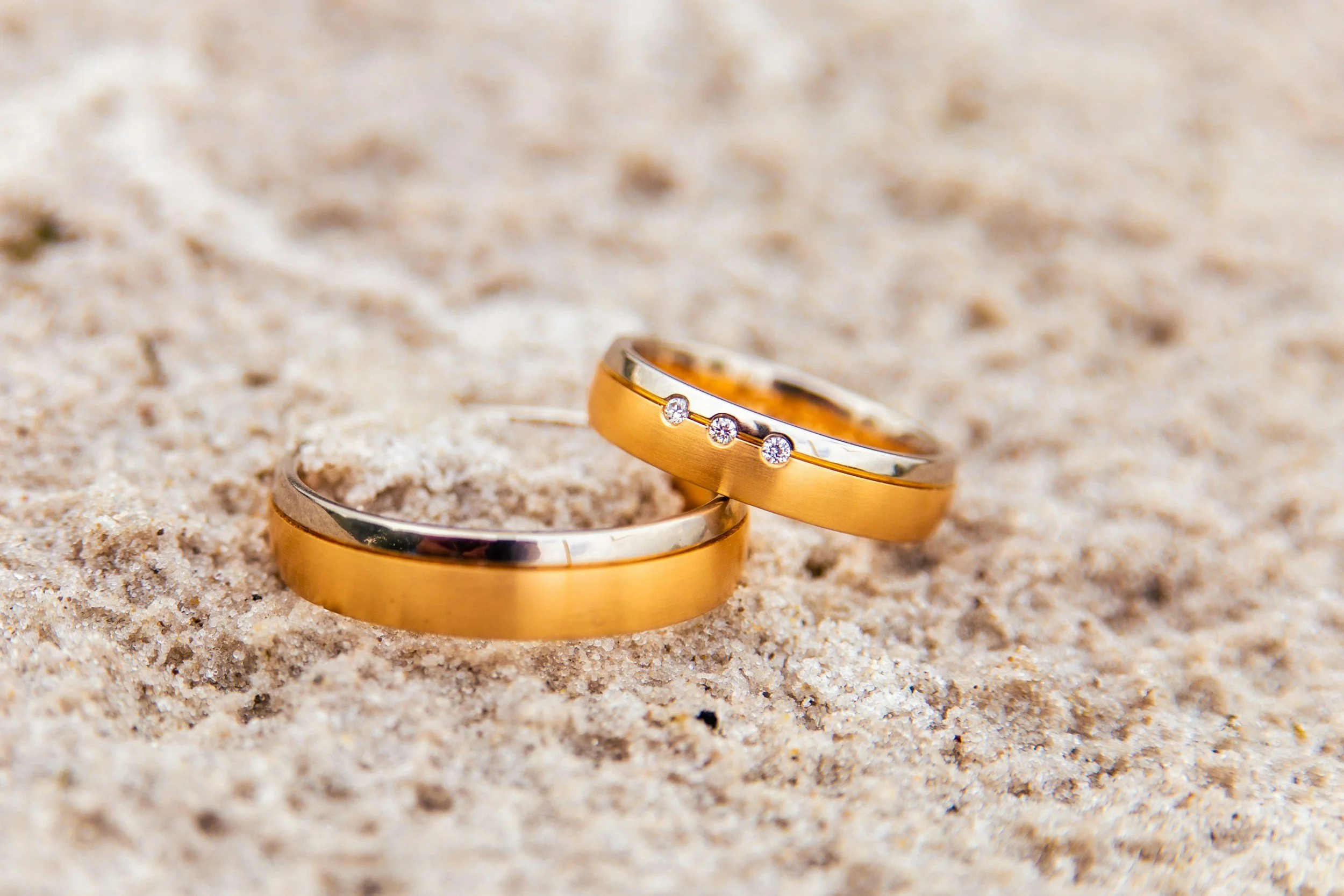 Two gold and silver wedding bands with one band decorated with three small diamonds, lying on sandy surface.