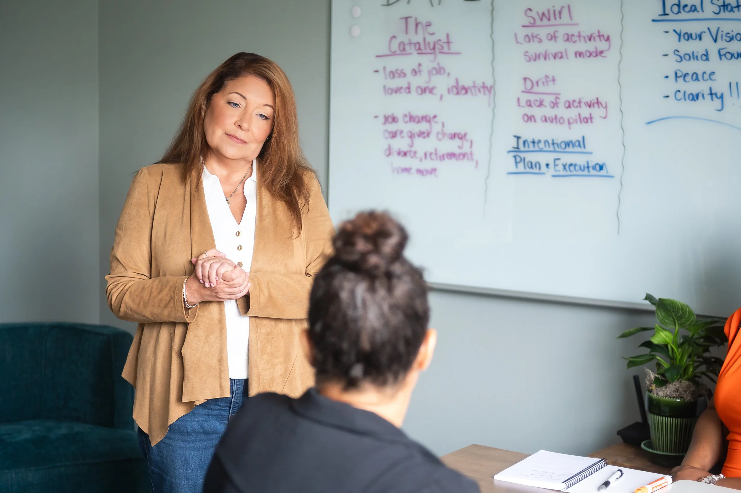 Patricia Rain, women's life coach, in her office looking sweetly at one of her clients. She has her hands folded, a soft smile, and is wearing a chestnut jacket, white button up and dark wash jeans.