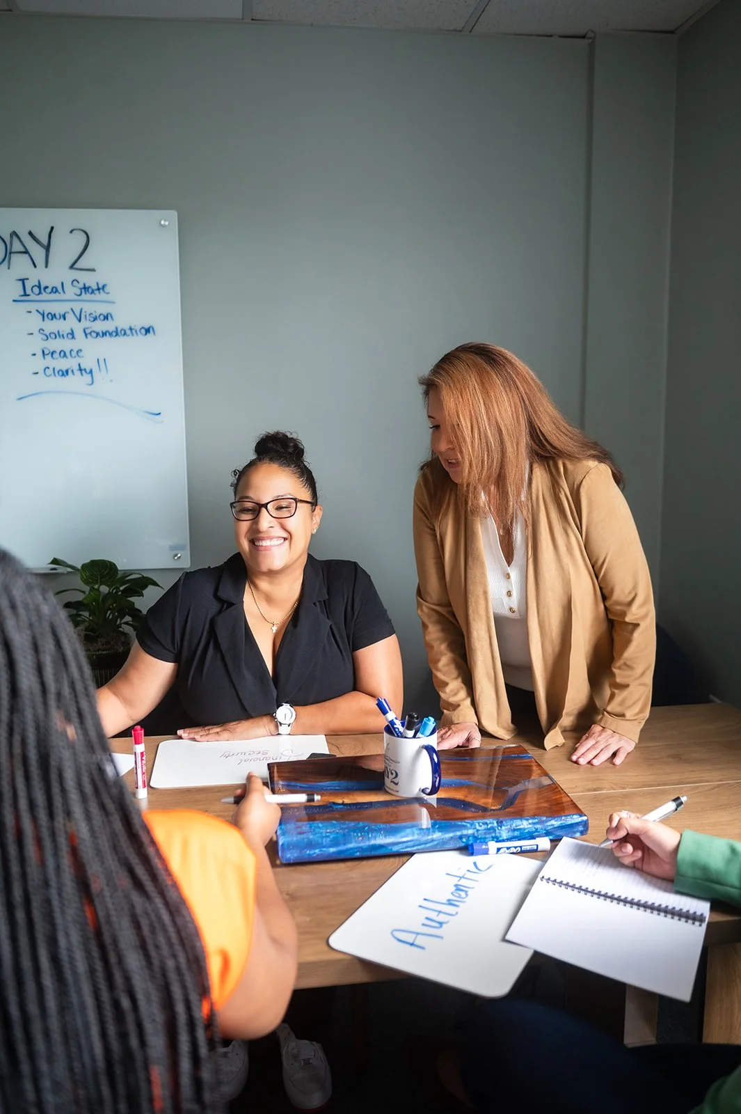 Patricia Rain, life and career coach, hosting a workshop in her Annapolis office. There is one client sitting down and smiling, the other chatting to her across the table, and Pat is standing above them looking at what they're writing on notebooks.