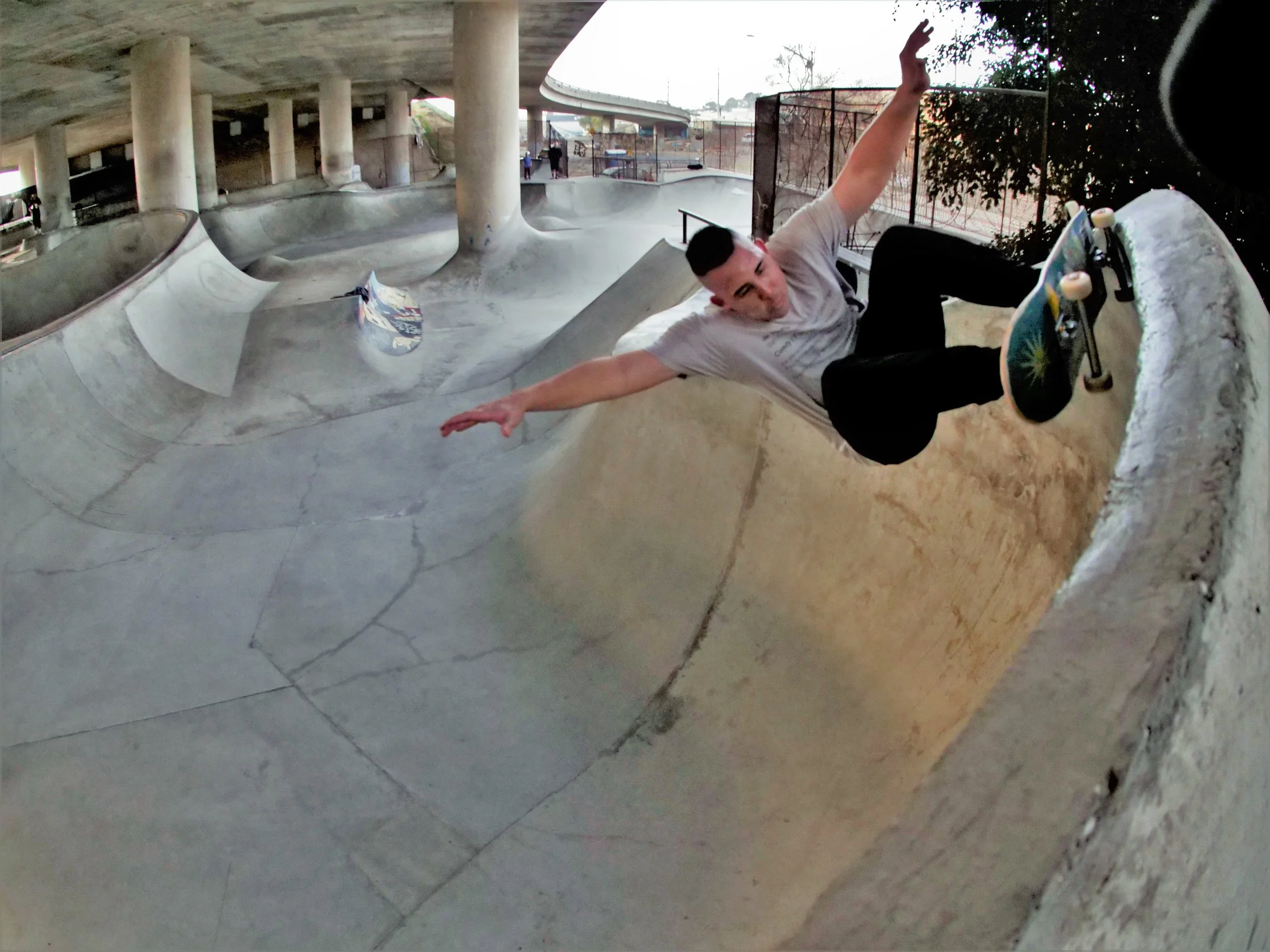 A skateboarder performing a trick at a concrete skate park under an overpass, viewed from a low angle with a skateboard in the foreground.