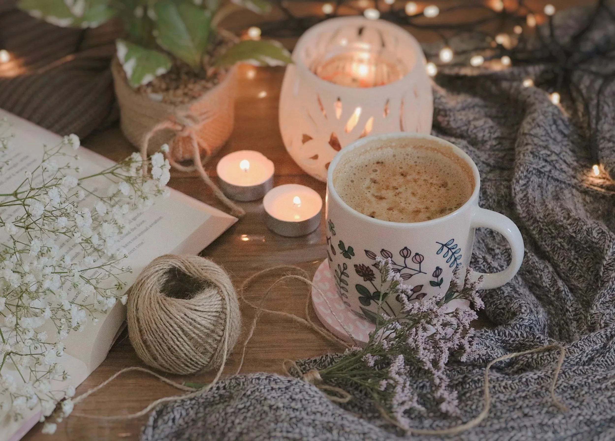 A cozy scene featuring a cup of frothy coffee, votive candles, a potted plant, a book with flowers, and decorative twine on a wooden surface, surrounded by fairy lights and a knitted blanket.