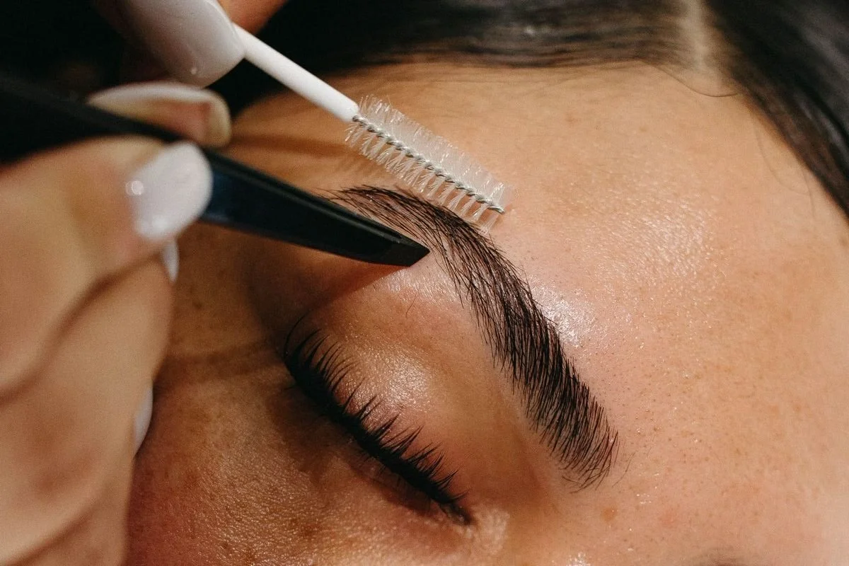 Close-up of a person receiving a brow lamination or tinting procedure, with a technician applying a treatment on the person's eyebrow using a small brush and tools.