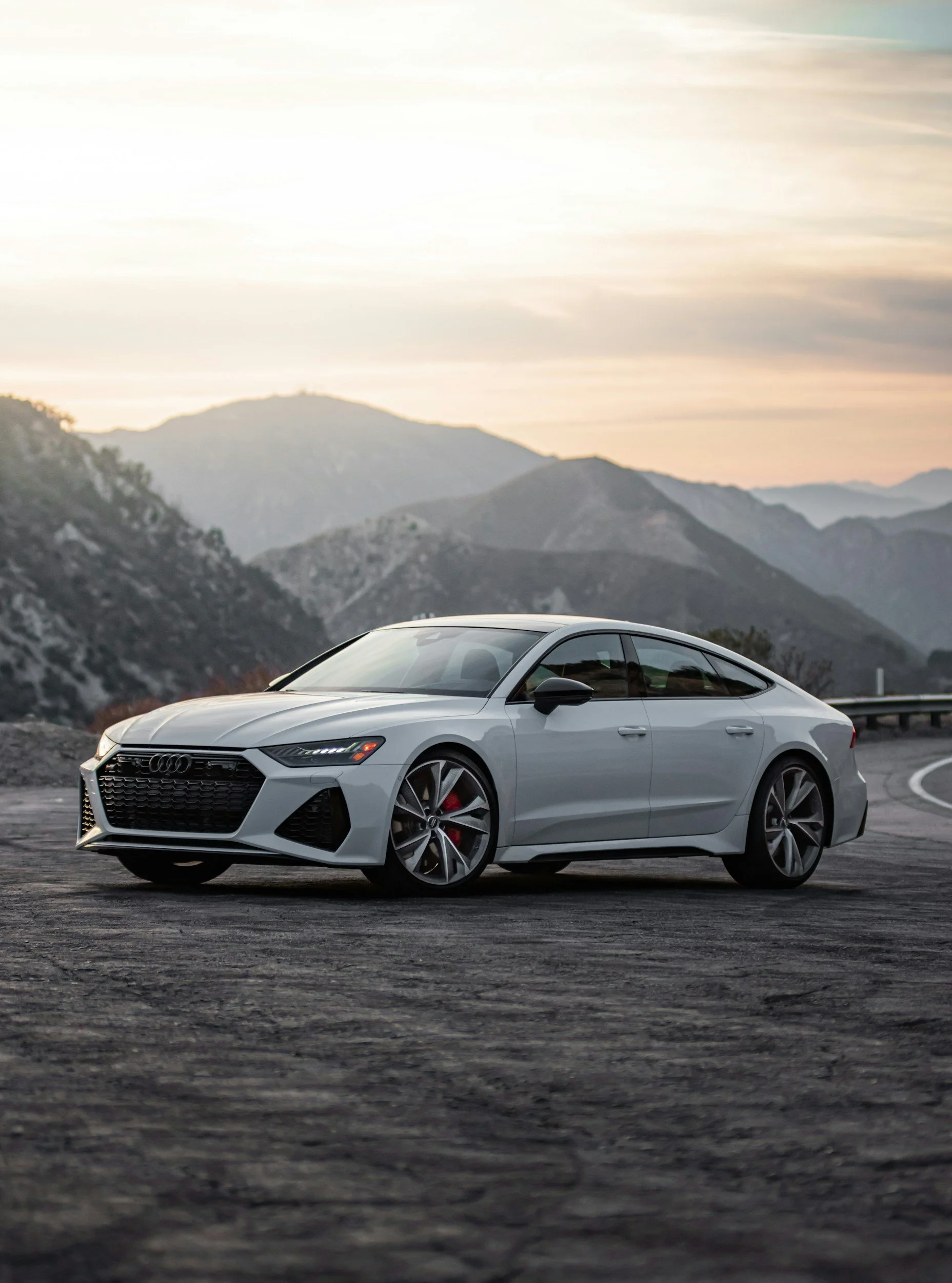 Gray Audi coupe car parked on a mountain road at sunset with mountains in the background.