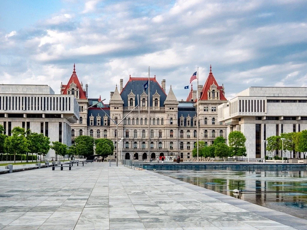 A historic-style courthouse with a gray stone facade, towers with red roofs, and American flags in front of a city plaza with trees and water features.