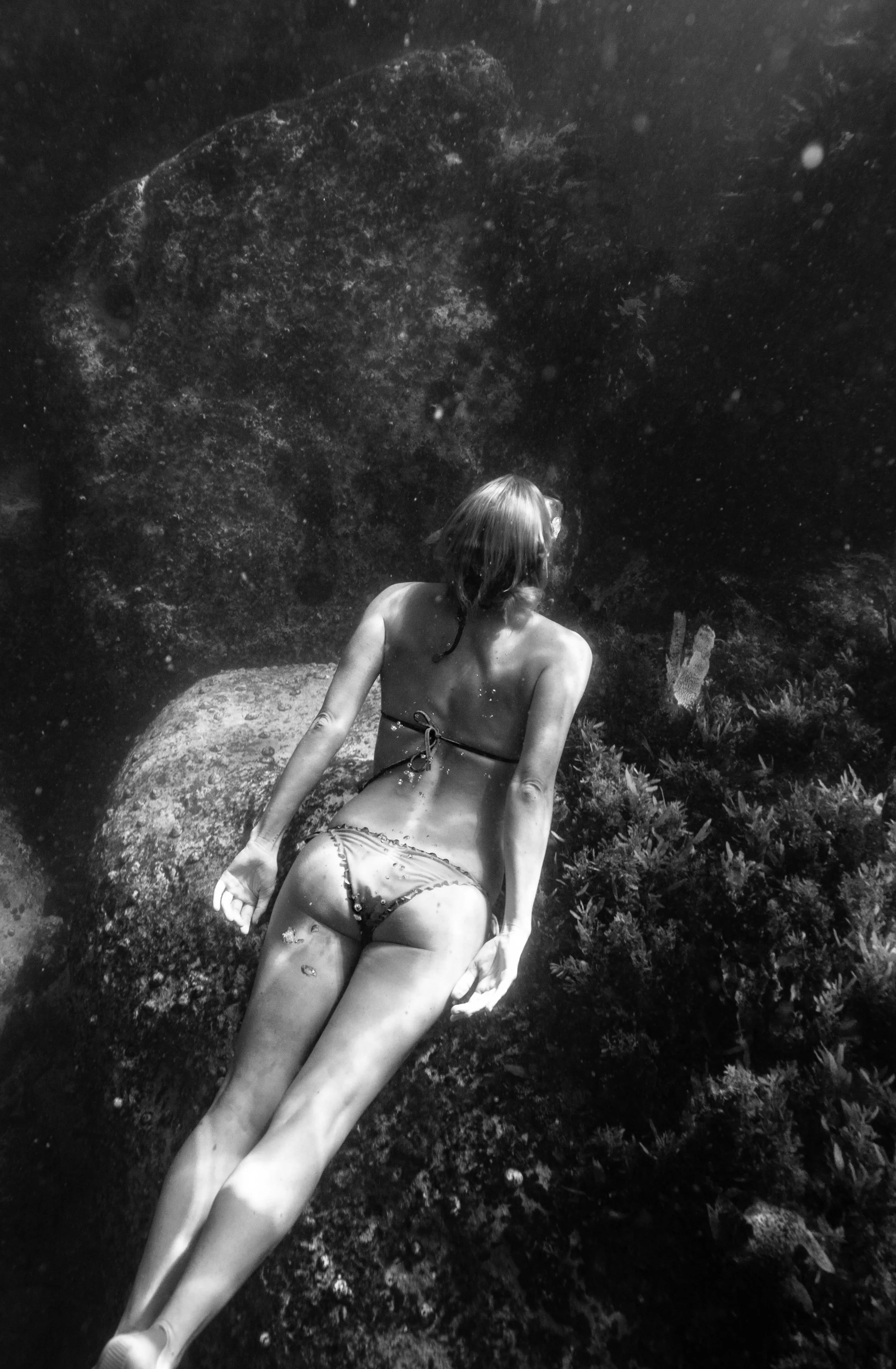A woman in a bikini sitting or lying on a rock underwater, with her back to the camera, in a natural setting with plants and rocks.