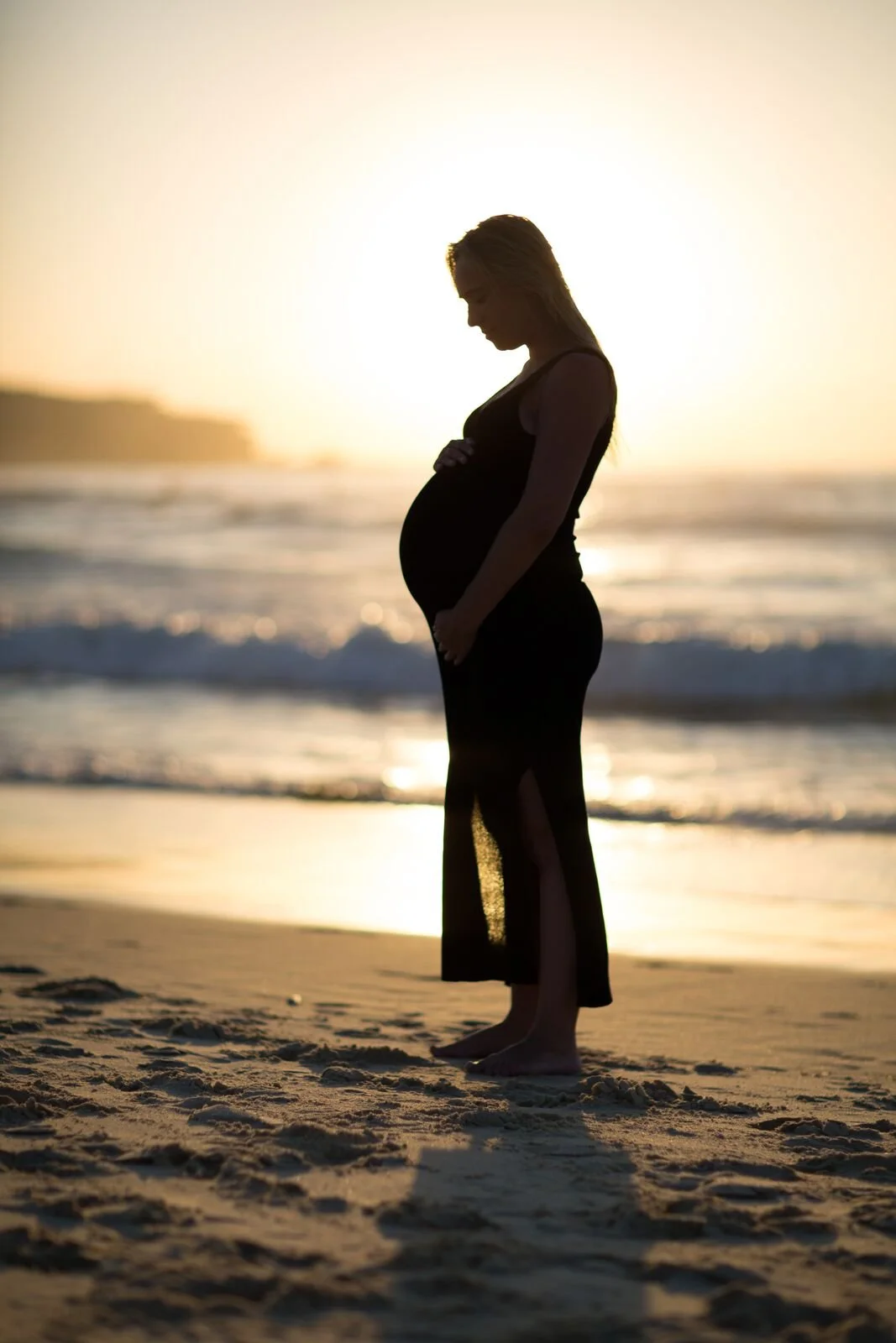 Silhouette of a pregnant woman standing barefoot on the beach at sunset, with her hands on her belly and head bowed in reflection.  Maternity style in Bondi, Sydney.