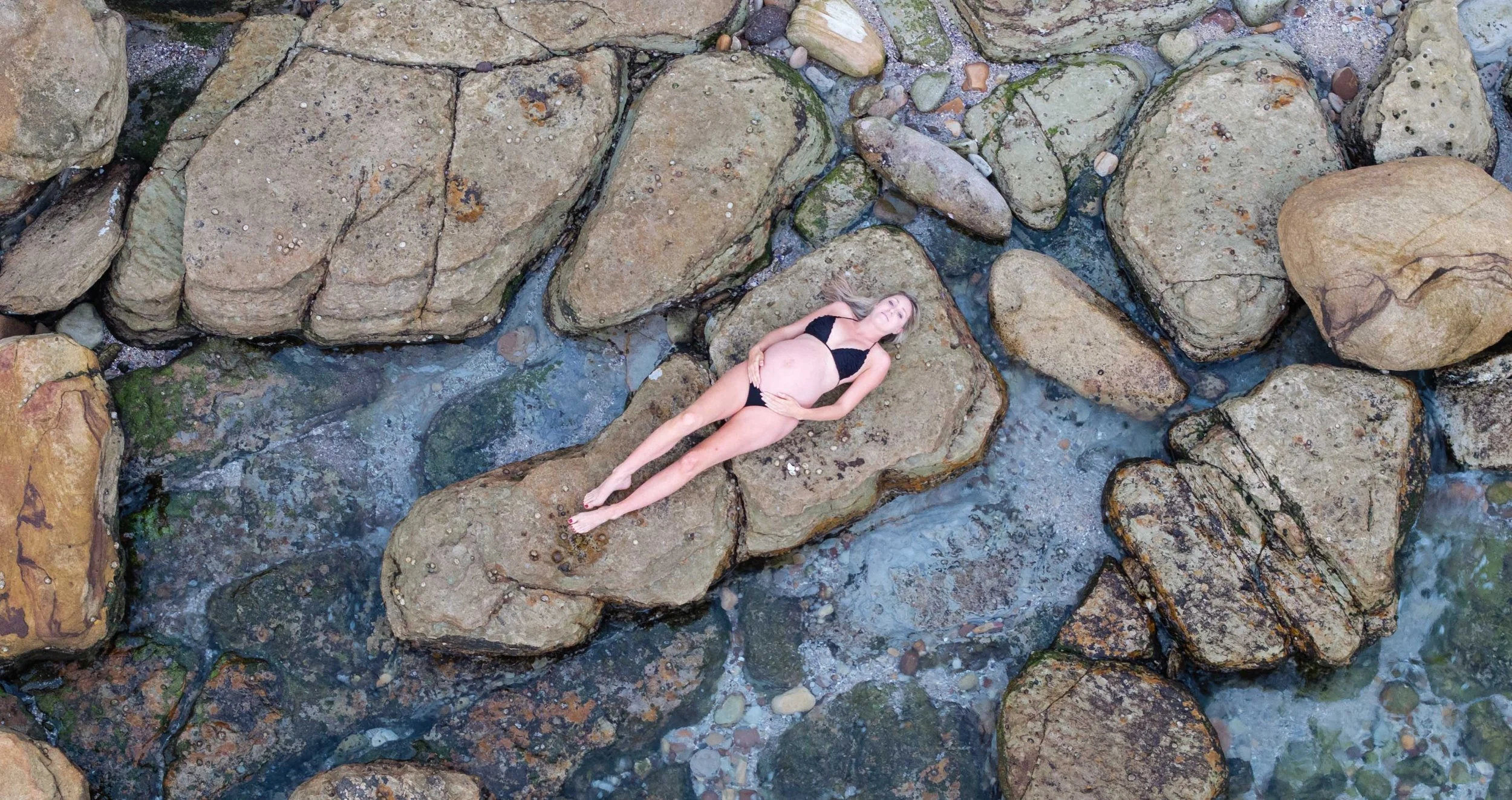 A pregnant woman lying on large rocks in a shallow stream, wearing a black bikini, looking up with her hands on her stomach. Maternity style in Sydney.